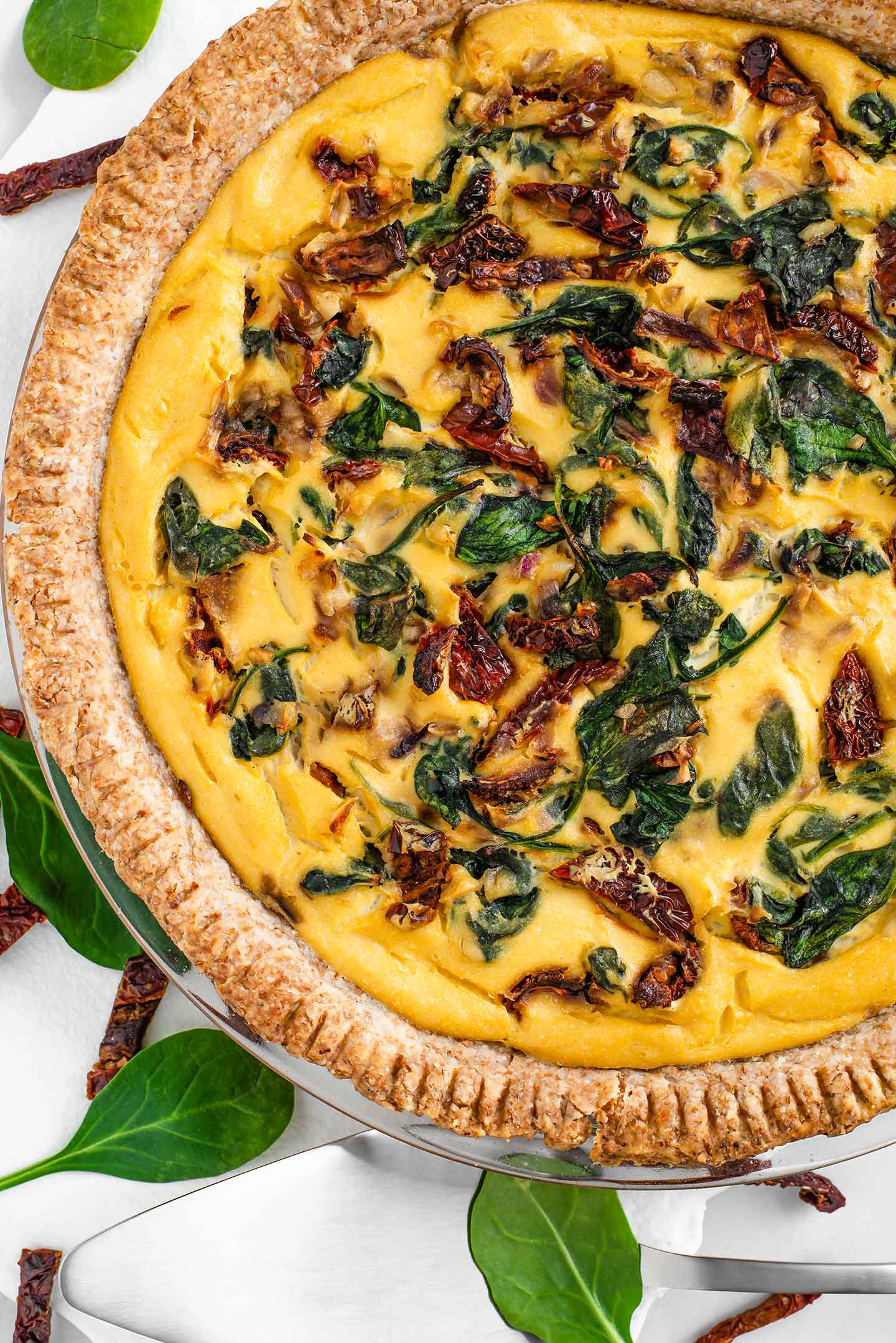 Top down view of a golden breakfast pie with sun-dried tomatoes and spinach. The edges of the whole wheat crust are crimped with a fork, browned, and crispy.
