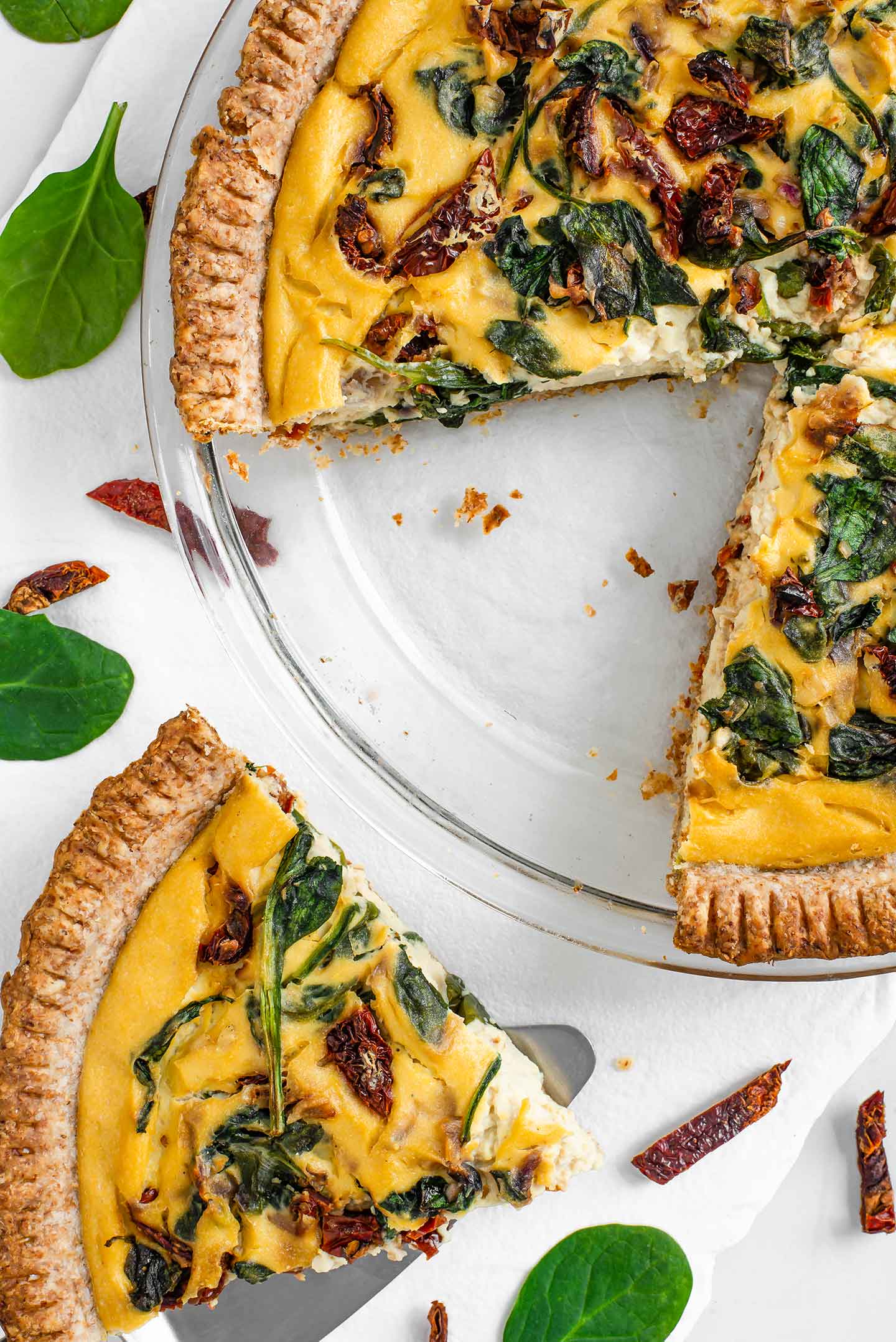 Top down view of simple spinach sun-dried tomato vegan "quiche" with a slice removed from the pie dish and placed on a small plate next to it.