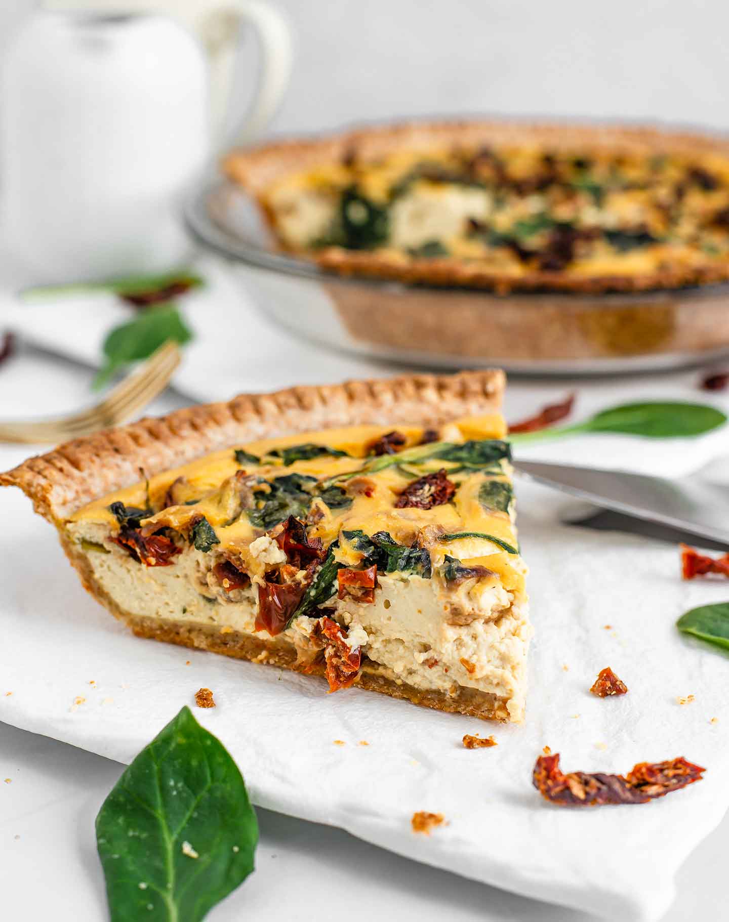 Side view of a slice of simple breakfast pie. The filling is thick and creamy with sun-dried tomatoes and spinach speckled throughout. The full pie with a piece missing is out of focus in the background.