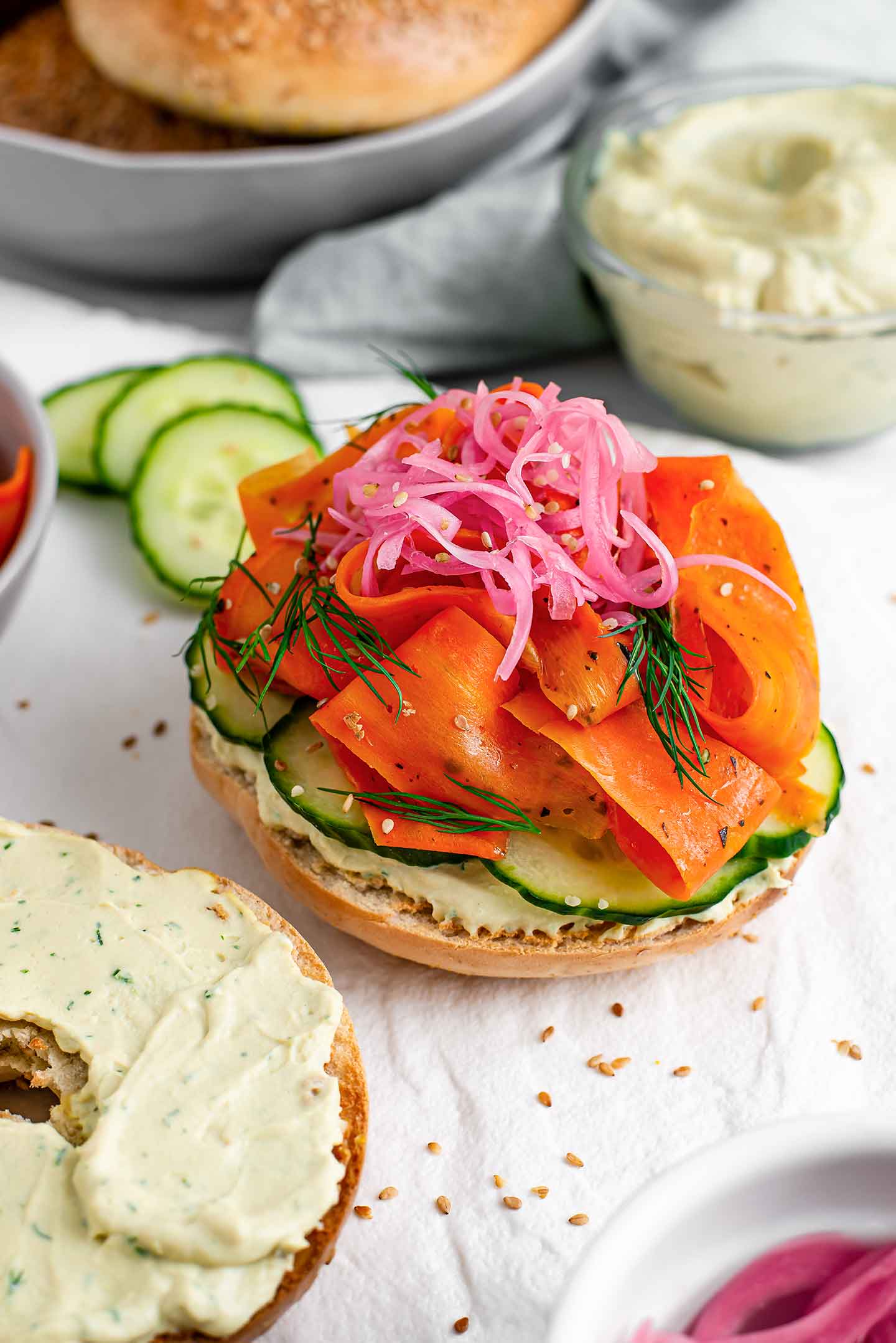 Close-up, top down view of a lox bagel sandwich without the top on. Vibrant pickled red onion and dill sit on top of smoky carrot lox, a bed of cucumber, and creamy dill spread.