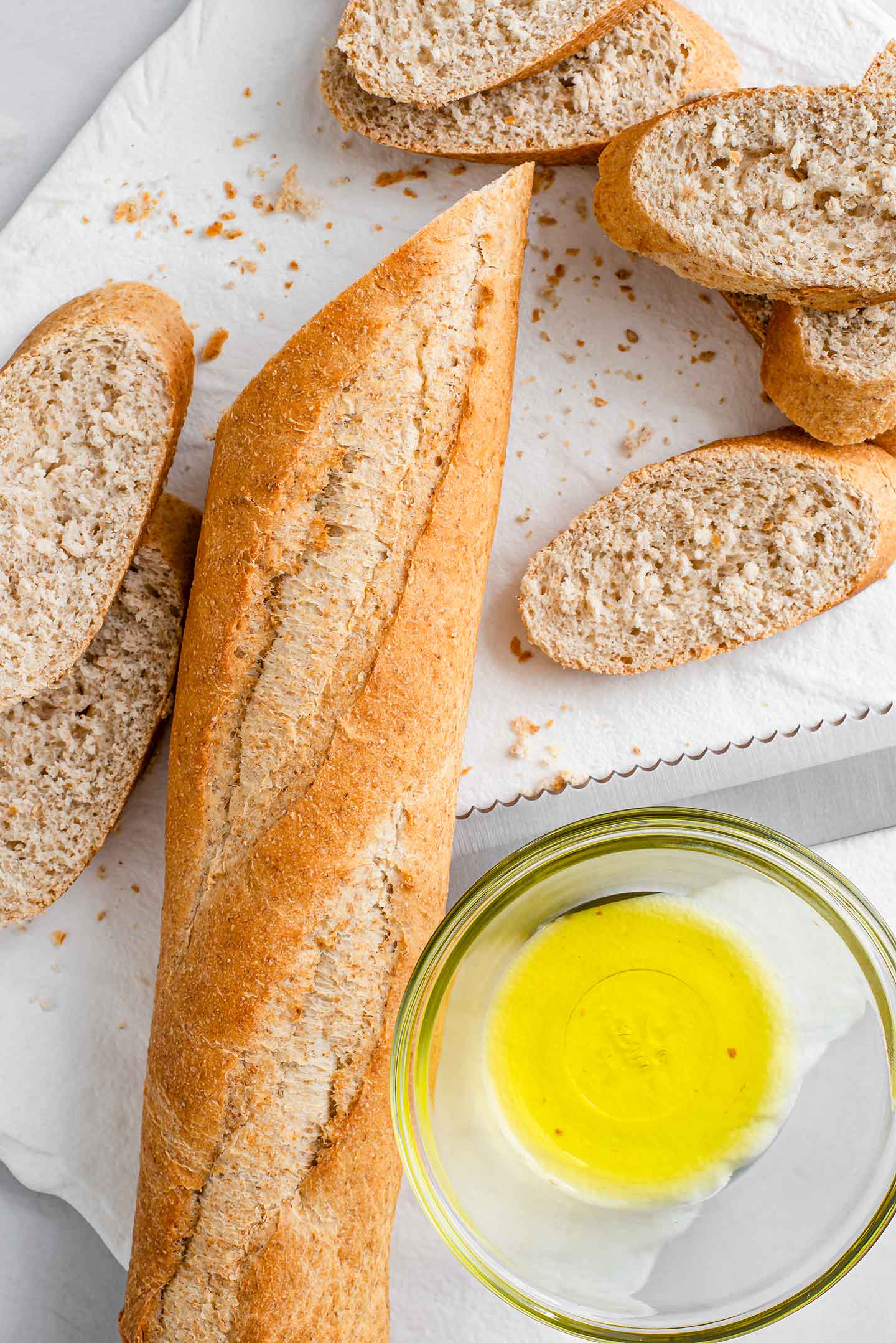 Top down view of a whole wheat baguette being sliced into thin crostini's. Olive oil is in a small glass bowl next to the bread.