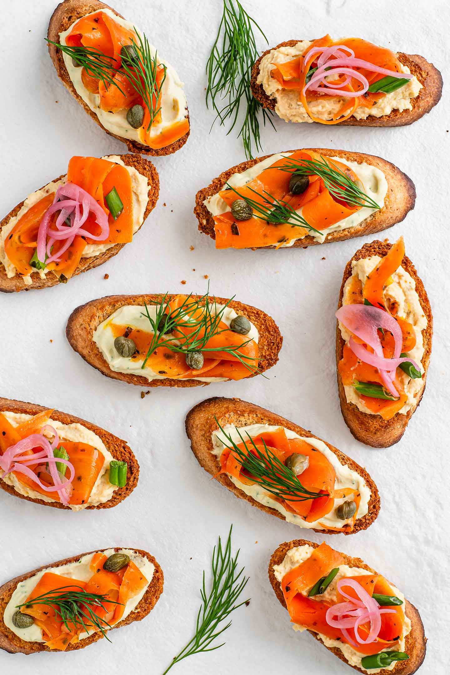 Top down view of smoky carrot lox appetizer displayed on a white tray. Crostini's are topped with dill spread, hummus, carrot lox, dill, capers, and pickled onion.