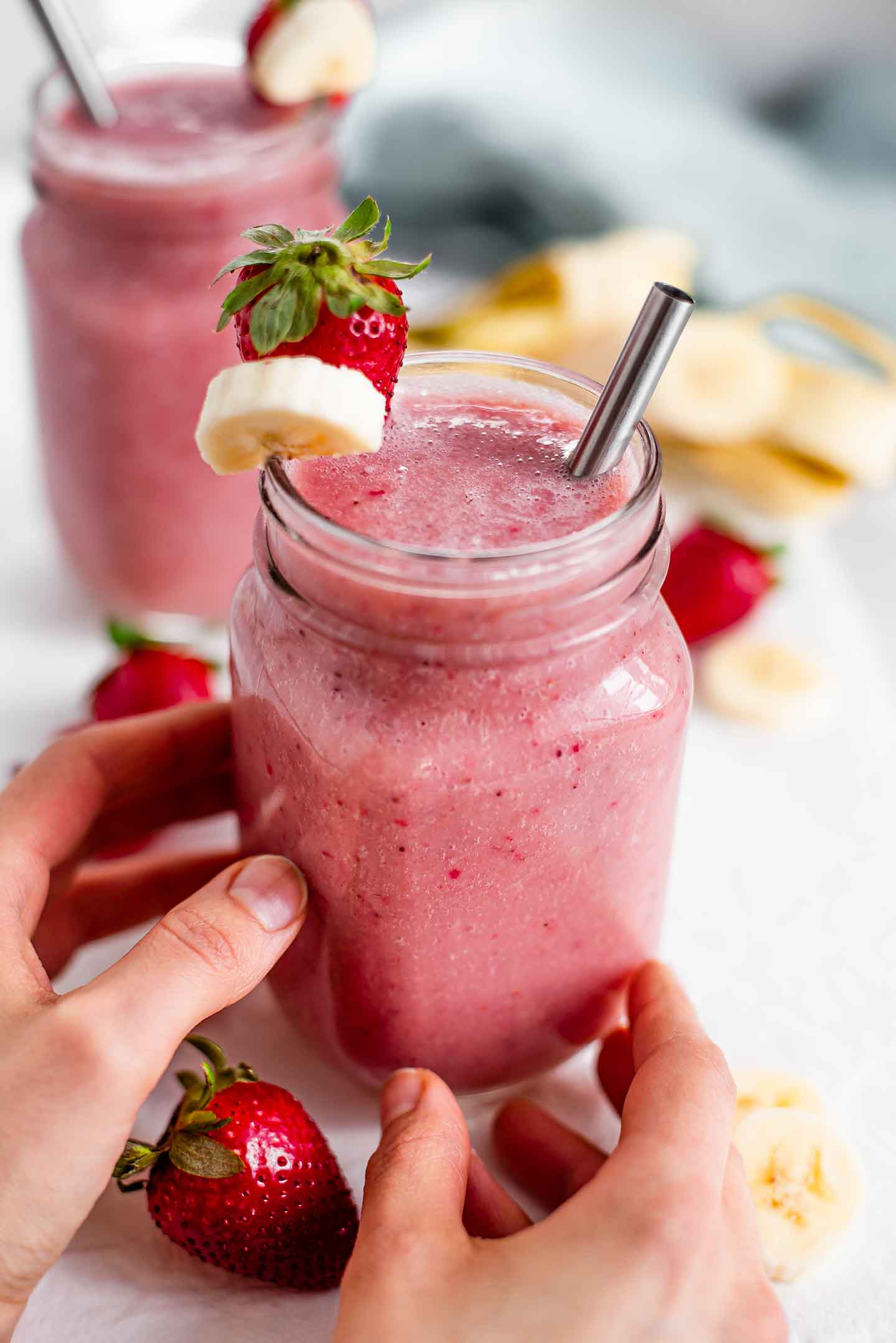 Side view of two hands reaching for a pink strawberry banana smoothie. A fresh strawberry and banana slice garnish the side of the glass jar and a stainless steel straw protrudes.