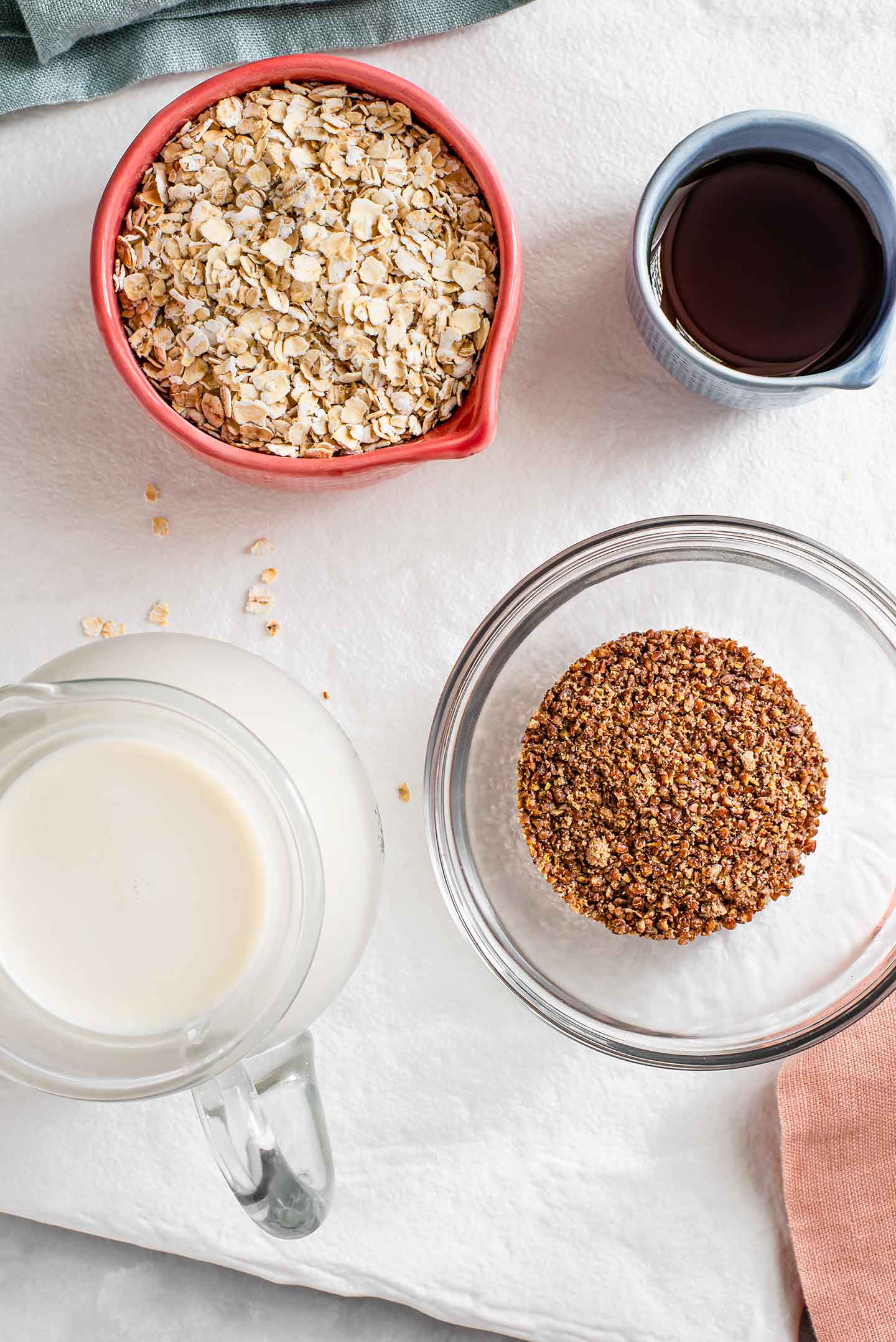 Top down view of ground flaxseeds in a small glass bowl with quick oats, maple syrup, and a small carafe of milk also displayed on a white tray.