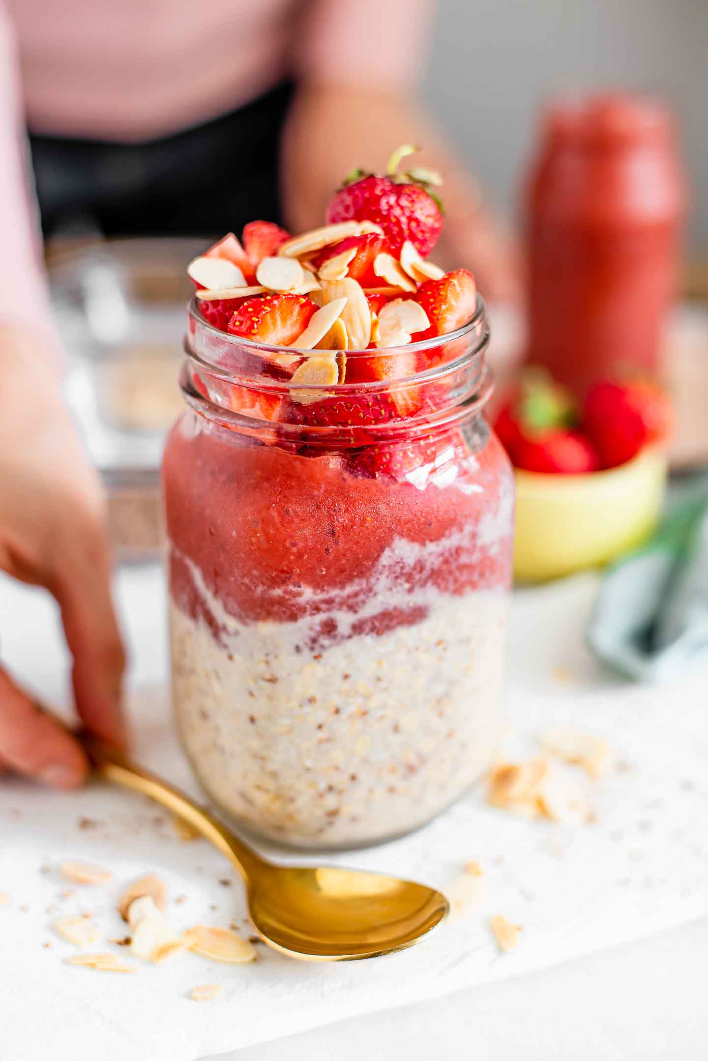Side view of a mason jar filled halfway with creamy overnight oats, then a layer of thick strawberry jam, topped with diced fresh strawberries and toasted sliced almonds. A hand picks up a gold spoon laying next to the jar.