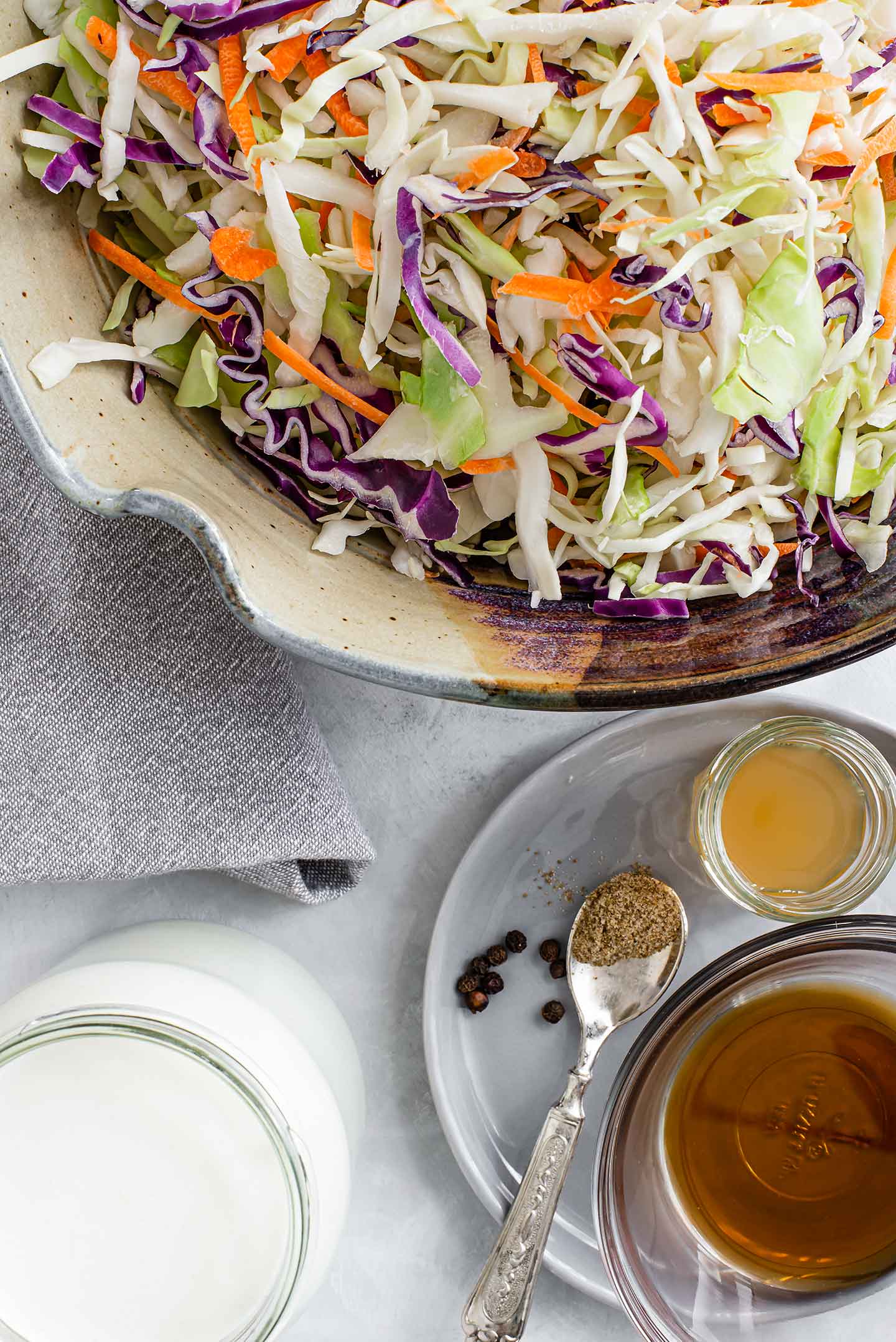 Top down view of ingredients for this summery vegan coleslaw recipe. Coleslaw mix, aquafaba mayo, maple syrup, apple cider vinegar, celery salt, and black pepper are displayed.