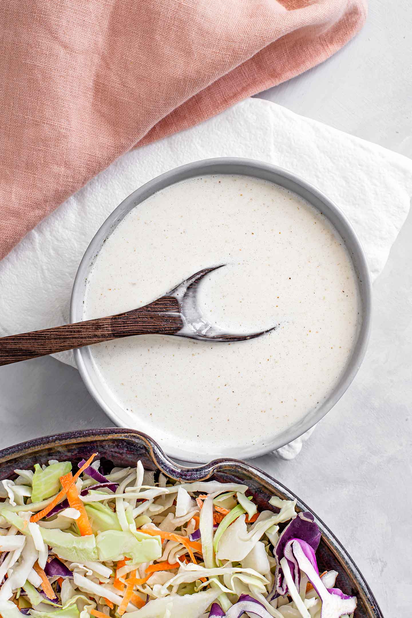 Top down view of vegan coleslaw dressing in a small dish with a spoon resting inside. The aquafaba mayo is speckled with celery salt and black pepper.
