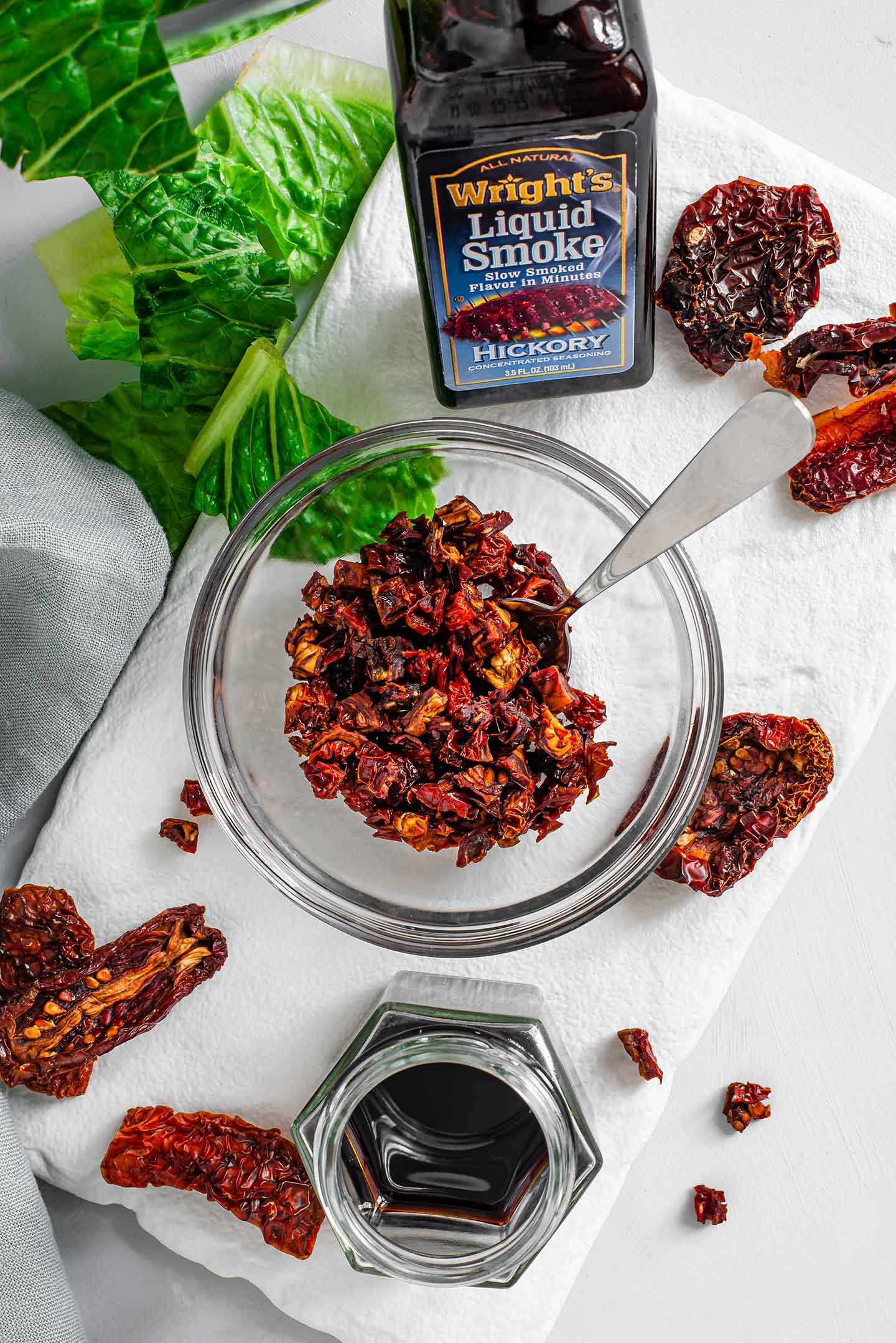 Top down view of sun-dried tomato bits in a small glass bowl with a bottle of liquid smoke, a jar of tamari, and scattered sun-dried tomato slices on a white tray.