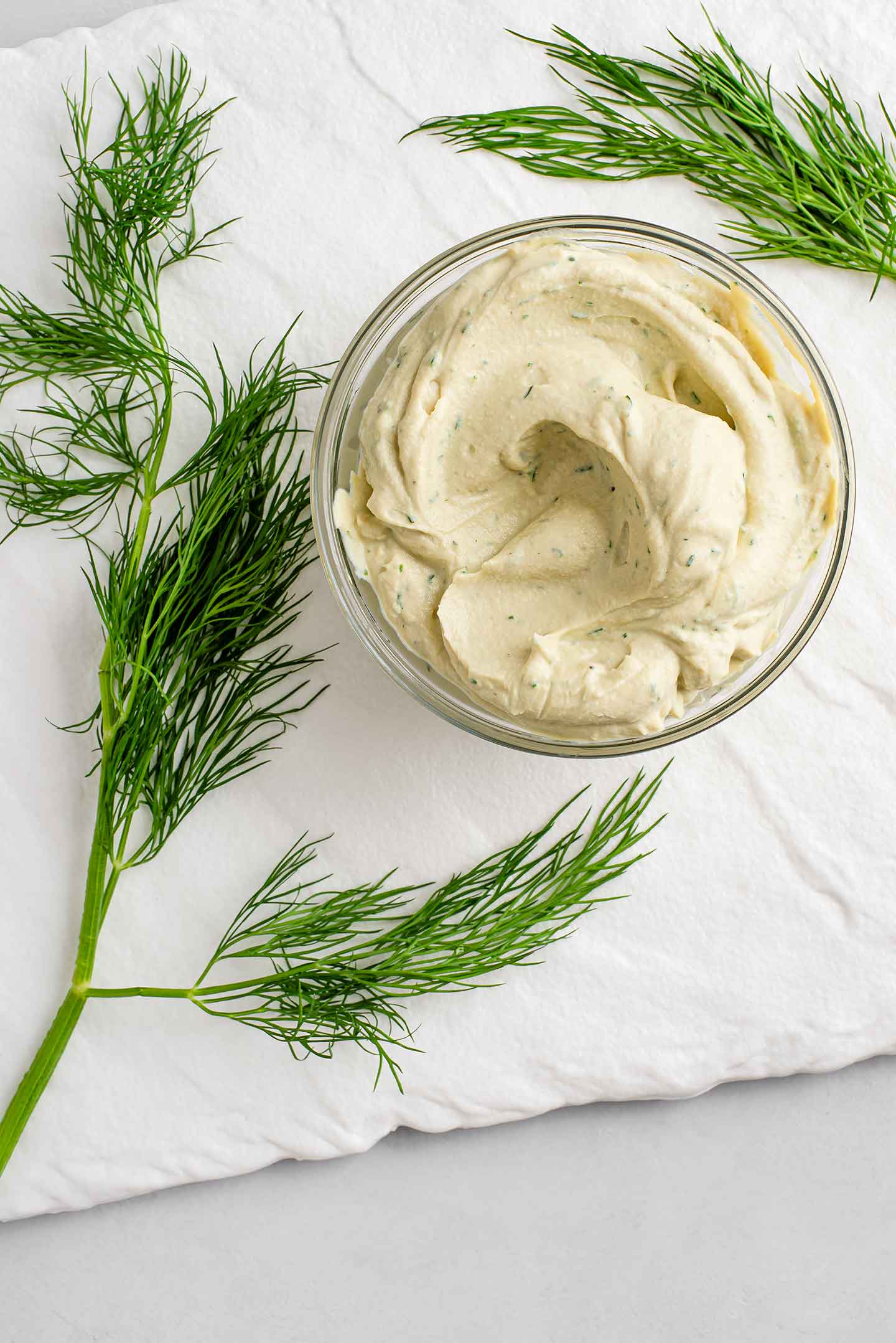 Top down view of vegan cream cheese with flecks of dill in a small glass dish on a white tray. Sprigs of fresh dill surround the dish.