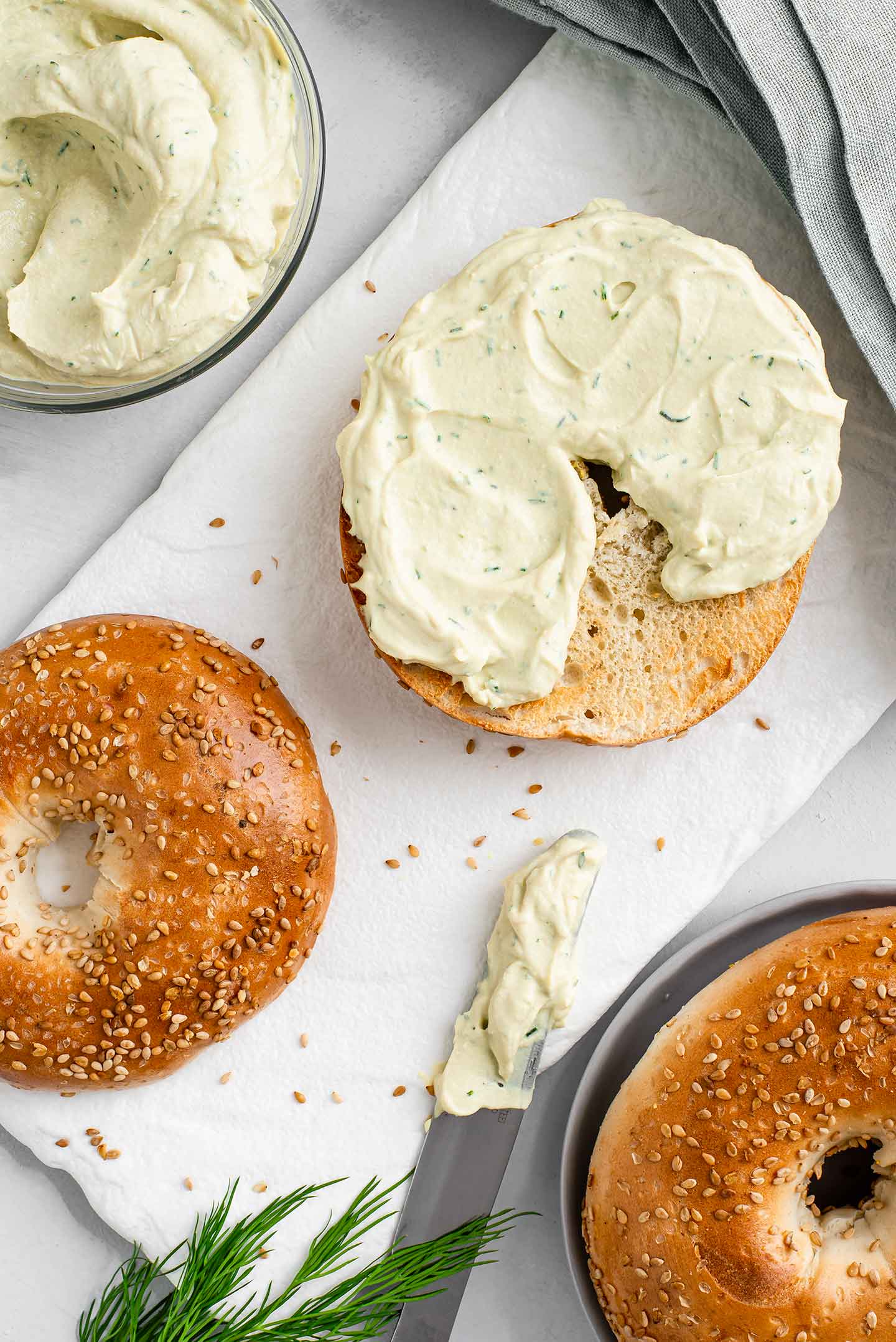 Top down view of creamy dill spread on half of a toasted sesame bagel. The other half lays on a tray next to it, a knife is covered in the creamy spread and more spread in a small bowl.