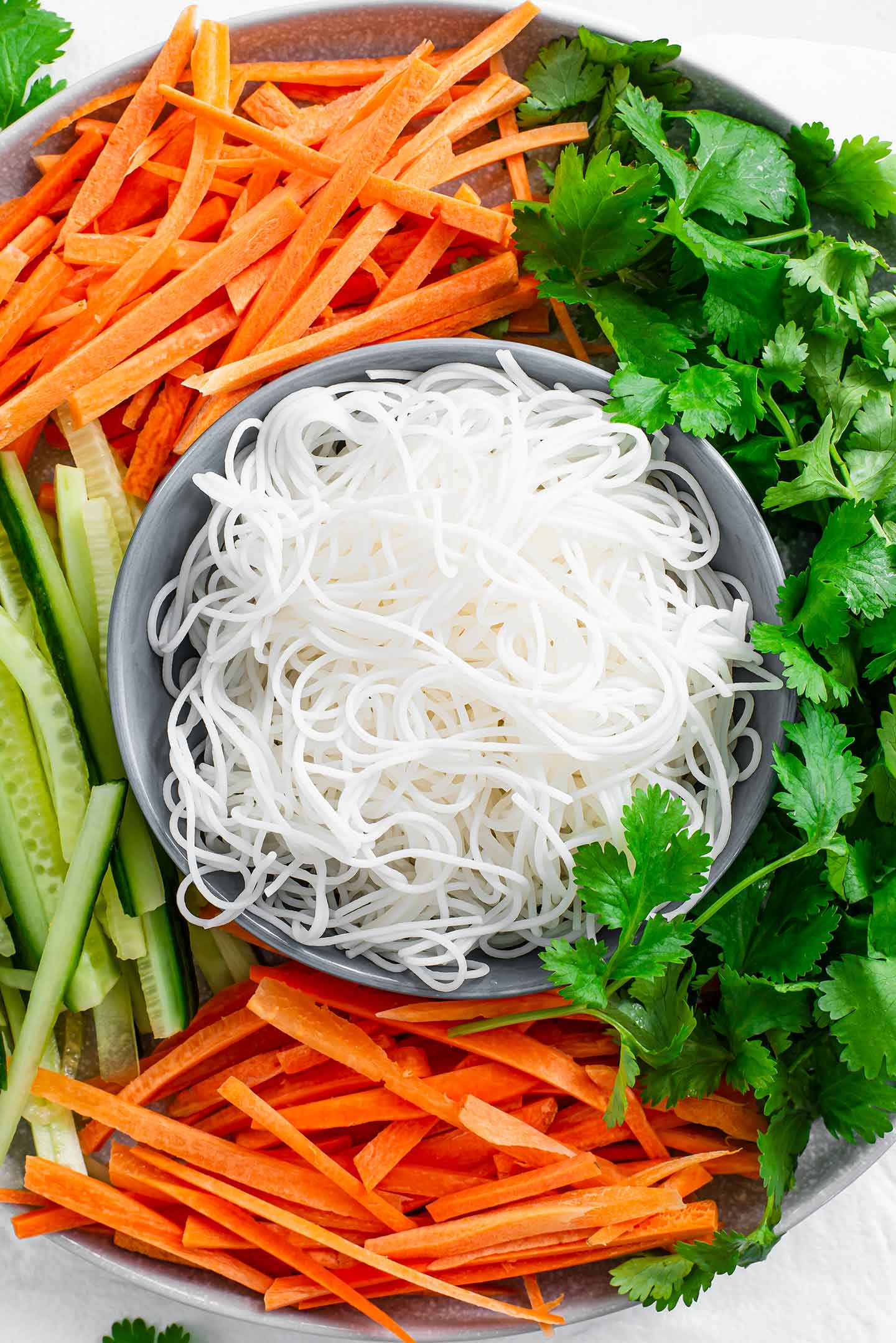 Top down view of ingredients for fresh spring rolls. Rice vermicelli noodles, cilantro, sliced carrots, and sliced cucumbers fill a plate.