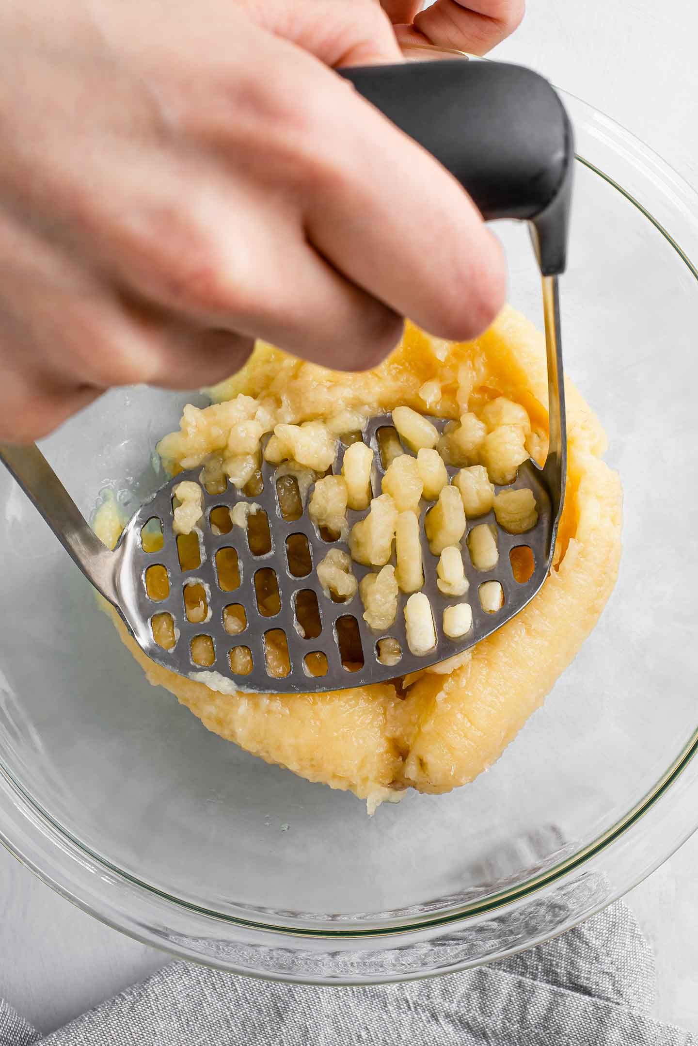 Top down view of a hand using a masher to mash ripe bananas in a bowl.