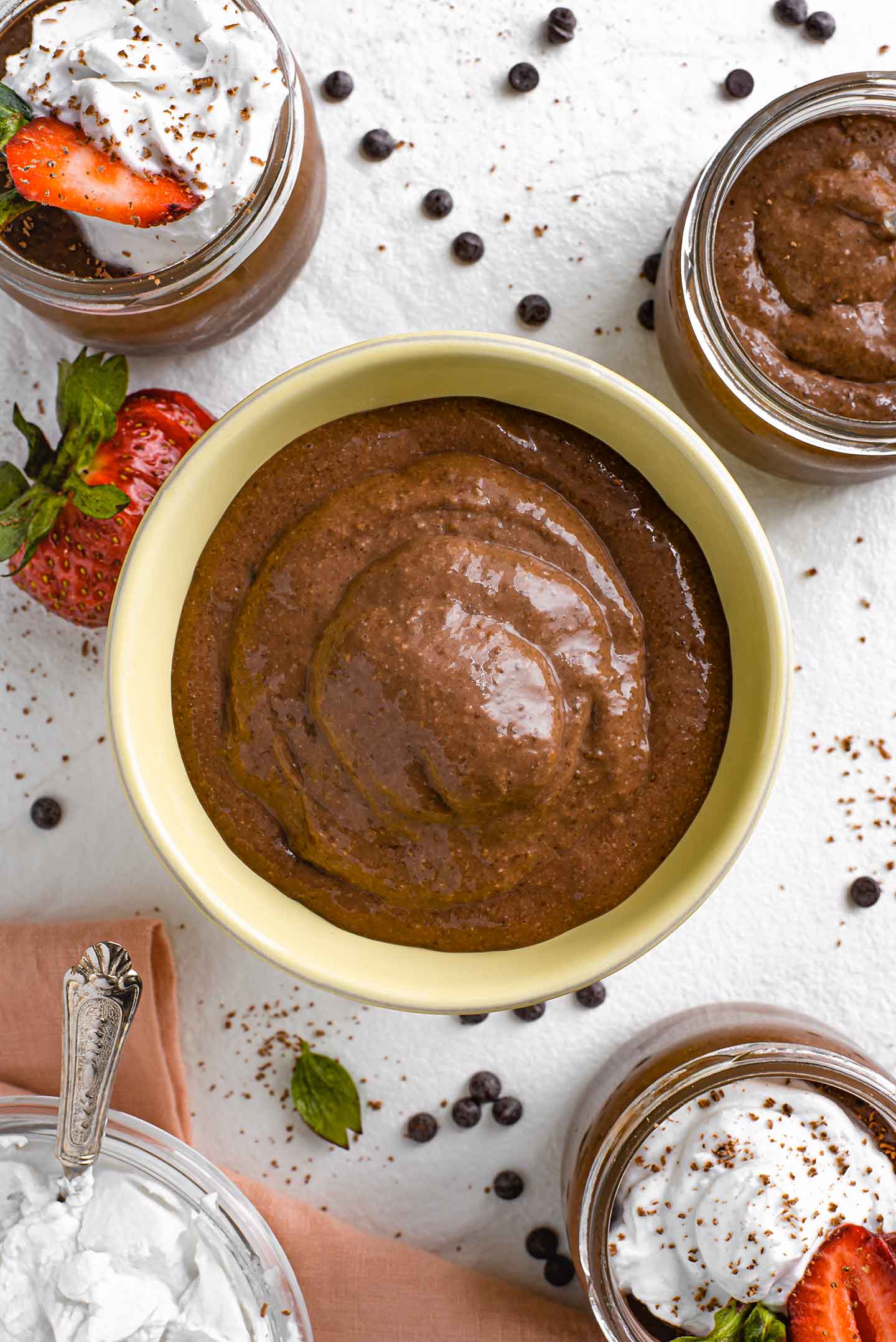 Top down view of thick chocolate pudding in a sorbet bowl with smaller 4oz portions in glass jars around it.