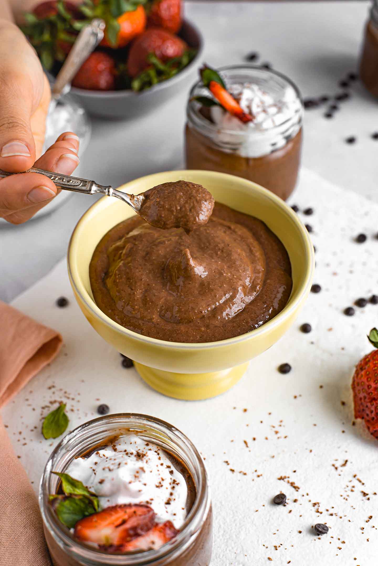 Side view of a spoon scooping thick chocolate pudding from a sorbet bowl. Chocolate chips and fresh strawberries are scattered around the bowl of pudding.