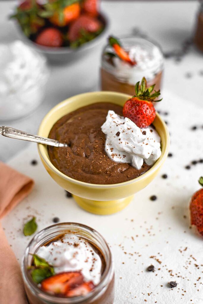 Side view of quick chocolate pudding filling a sorbet bowl with two 4 oz servings in glass jars around. Whipped cream and fresh strawberries garnish the pudding.