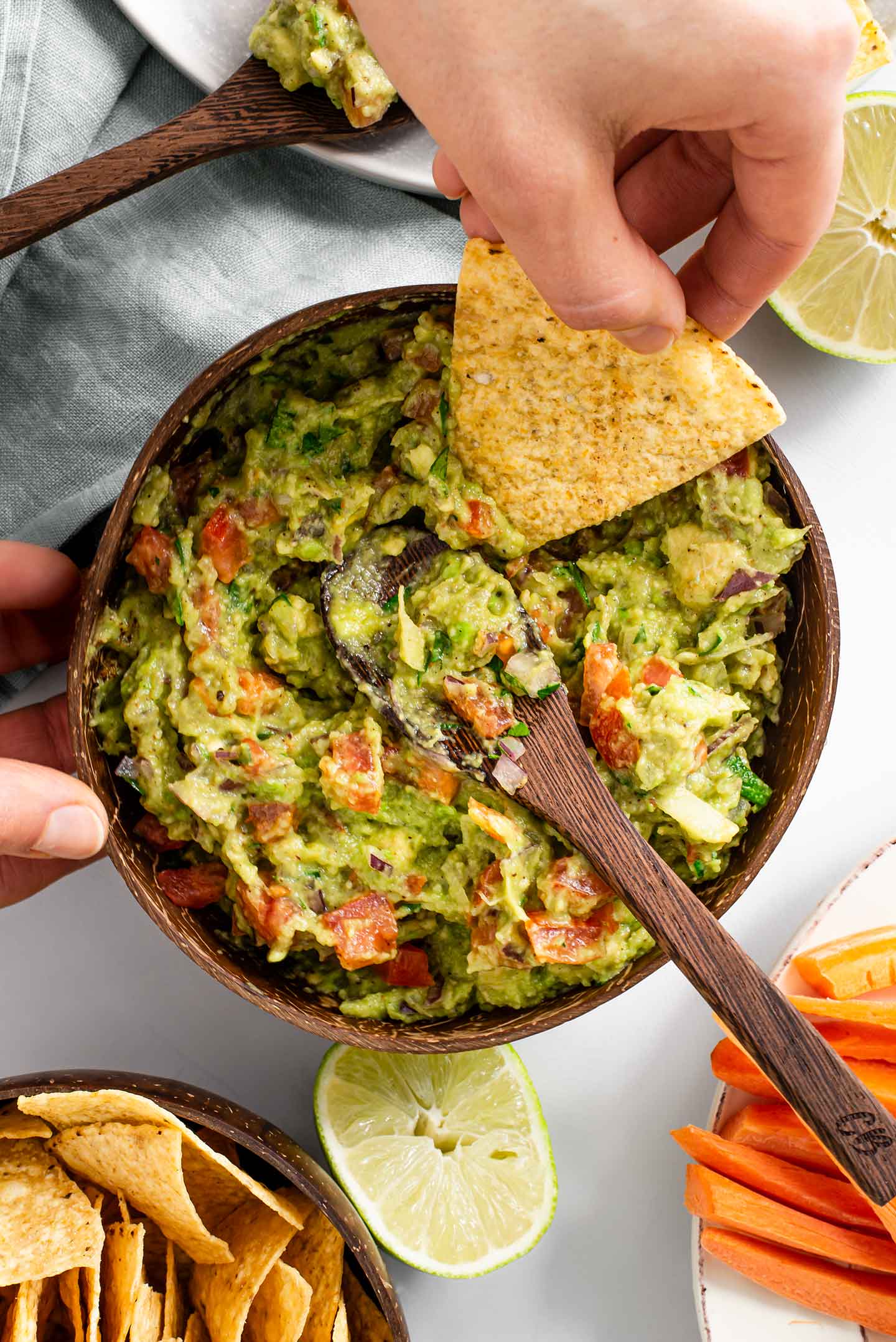 Top down view of a hand scooping timely guacamole from the bowl and onto a tortilla chip. Lime halves, carrot sticks, and more tortilla chips surround the guacamole.