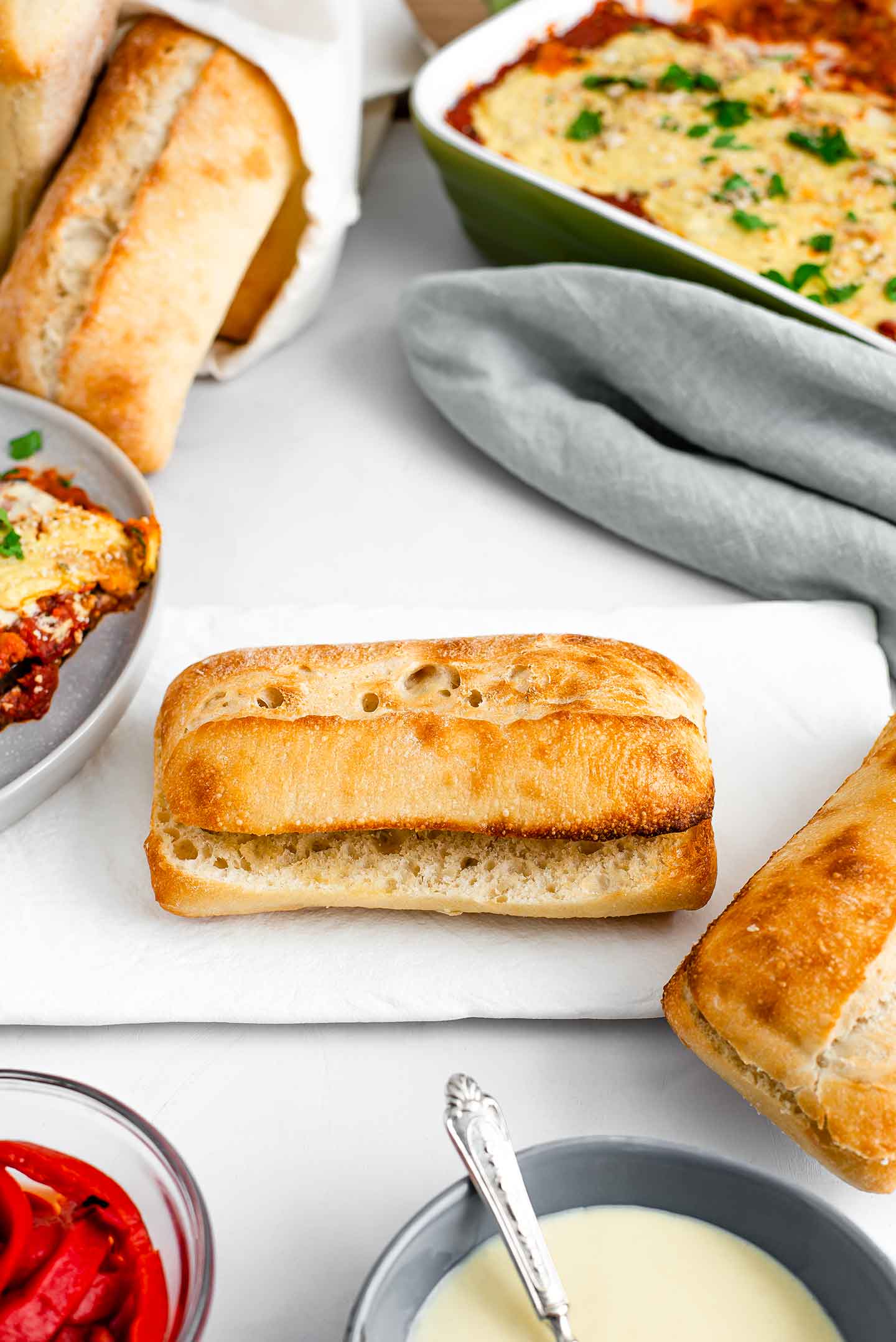A long ciabatta bun sits on a white tray with eggplant parm in a casserole dish, extra bechamel in a small bowl, and roasted red pepper in another small dish nearby.