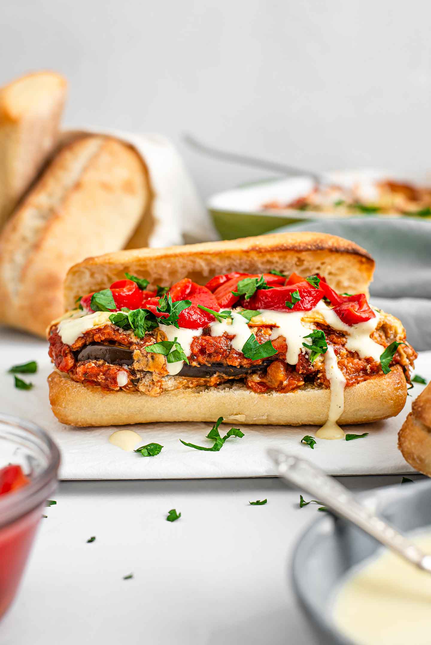 The top of a ciabatta bun rests against the loaded sandwich. Messy eggplant parmesan is topped with drizzled bechamel, parsley, and sliced roasted red peppers. More buns and eggplant parm are in the background.