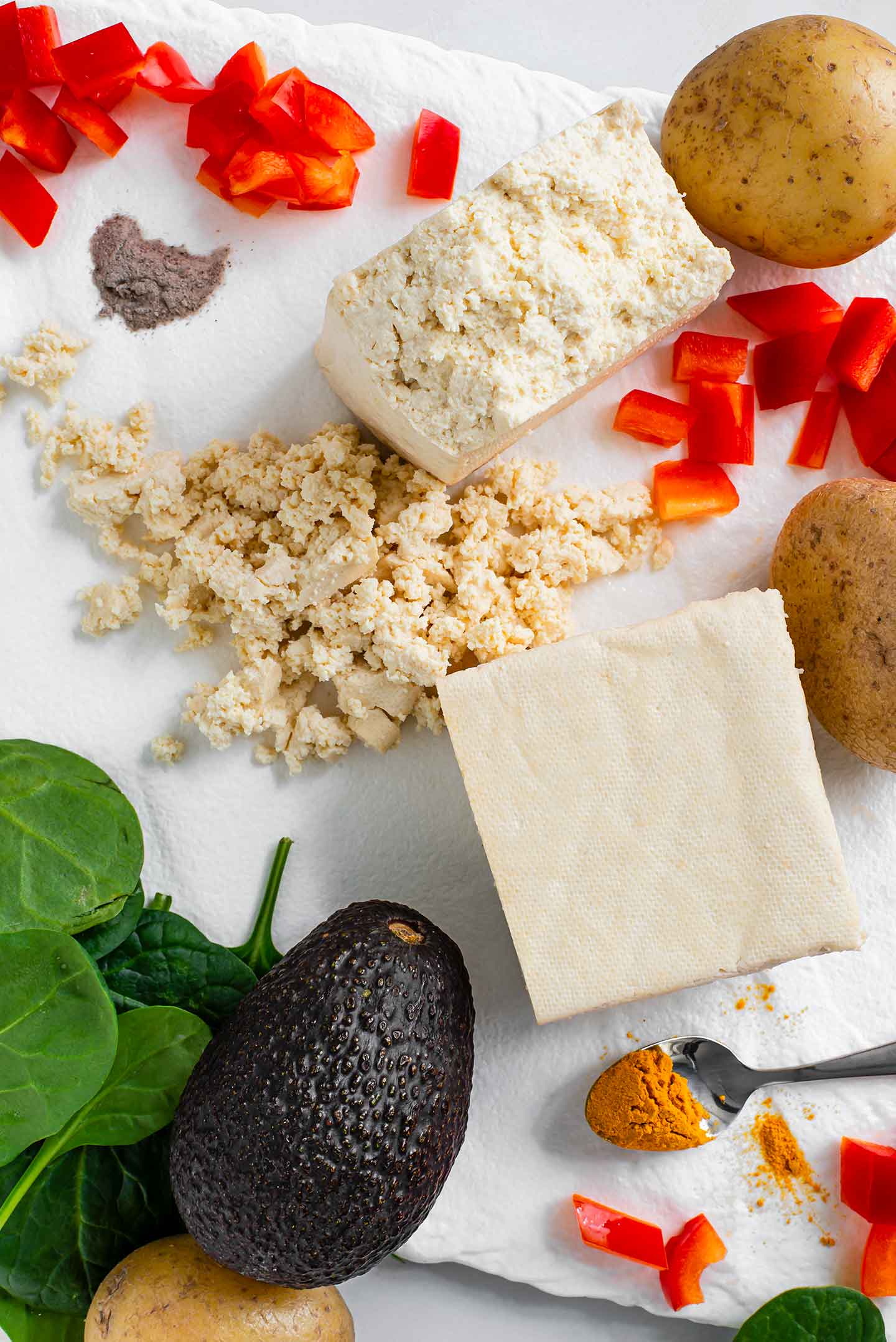 Top down view of ingredients for Scramble and Spuds Breakfast Bowl. Crumbled tofu, yukon gold potatoes, avocado, red onion, spinach, turmeric, and plate salt fill a white tray.