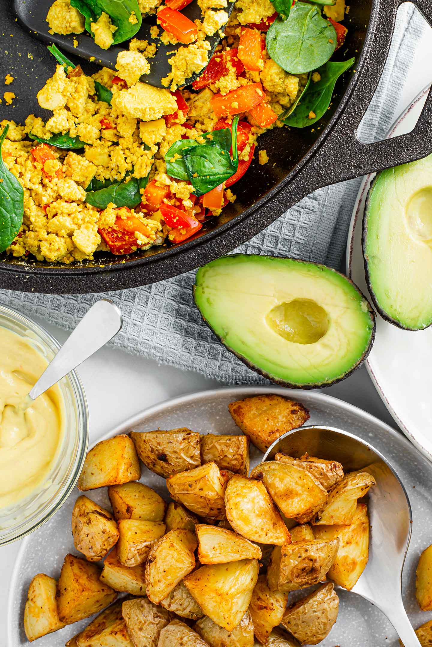Top down view of roasted potatoes in a bowl, tofu scramble in a cast-iron skillet, avocado halved, and cheesy queso in a small glass dish.