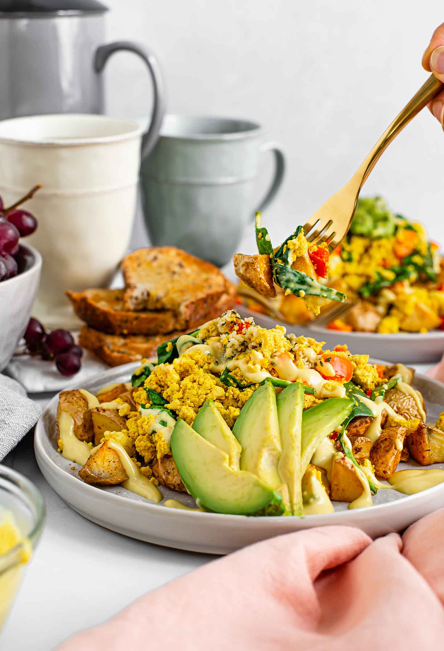 Side view of ultimate scramble and spuds breakfast bowl with sliced avocado and drizzled queso. Toast, grapes, coffee, and another scramble bowl are in the background.