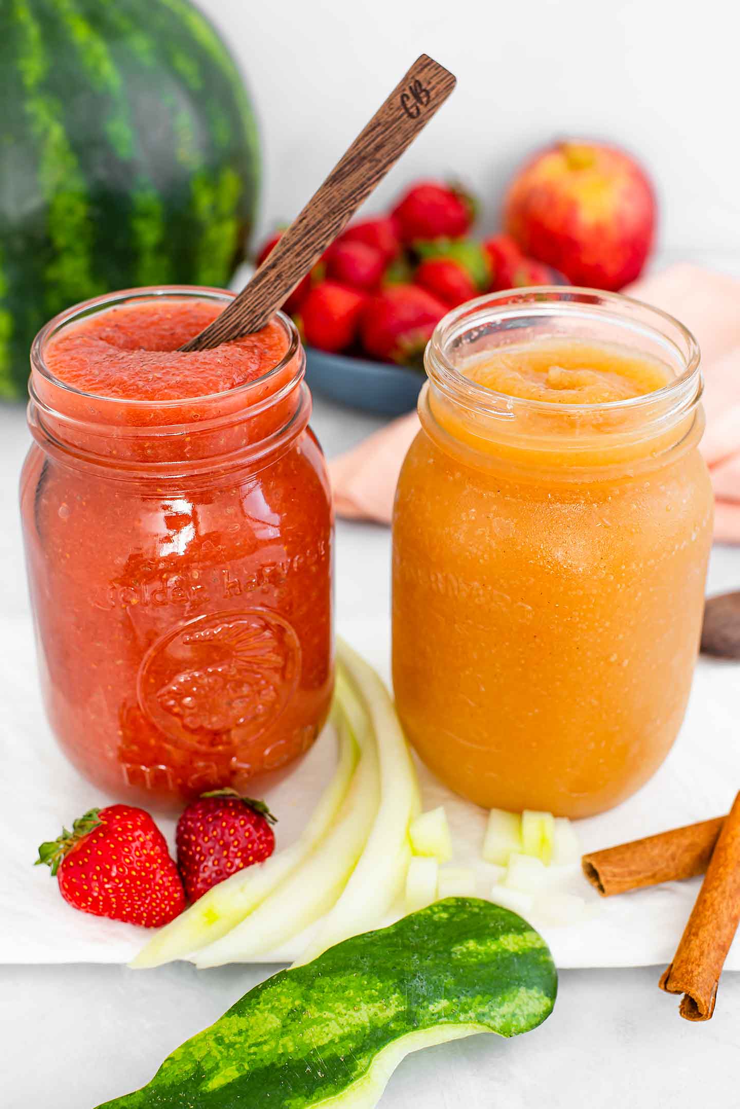 Side view of two jars of watermelon rind fruit sauce. A wooden spoon dips into a strawberry sauce and cinnamon sticks and an apple surround the golden watermelon applesauce.
