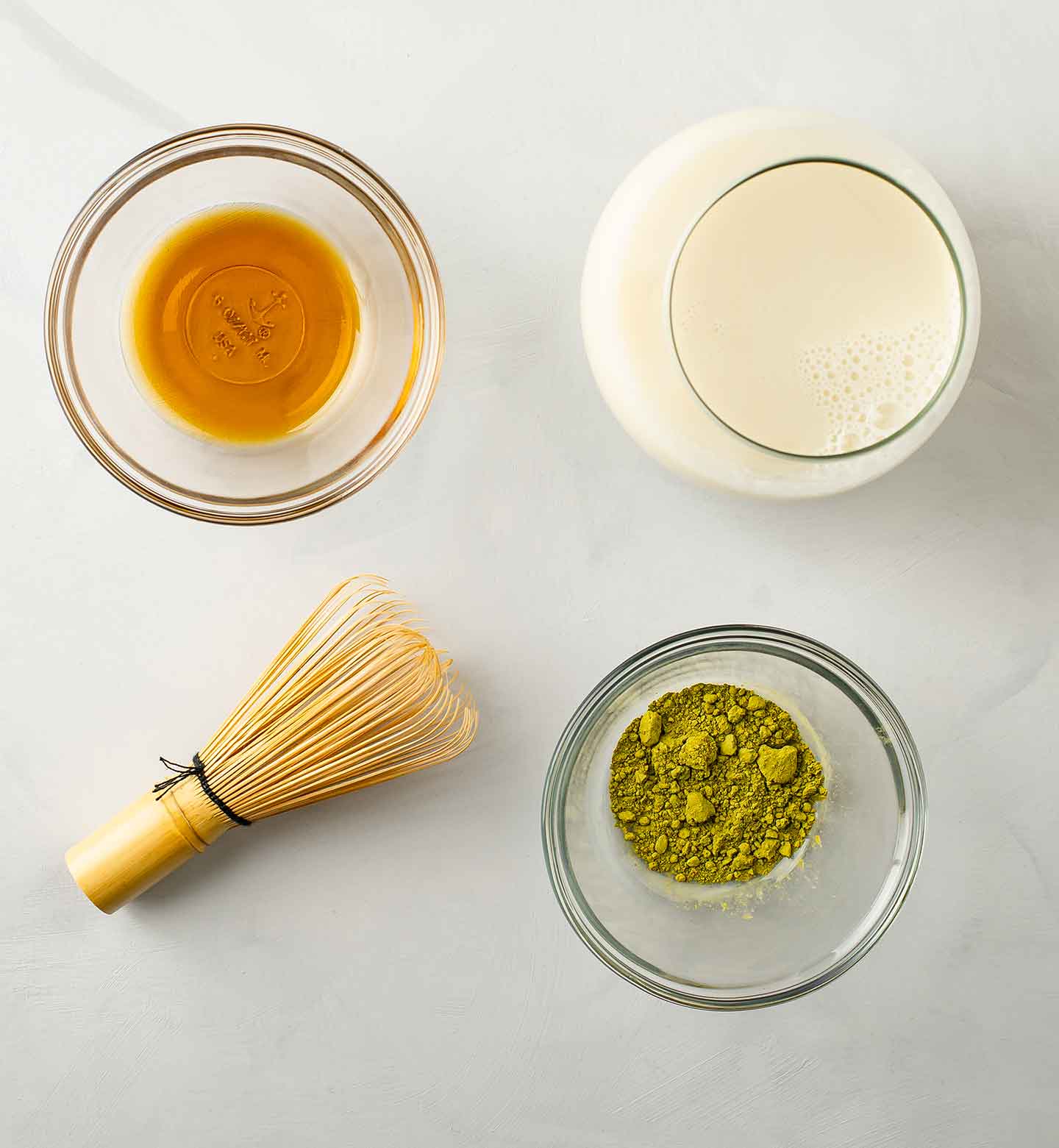 Top down view of a matcha whisk and matcha powder, milk, and maple syrup in separate bowls