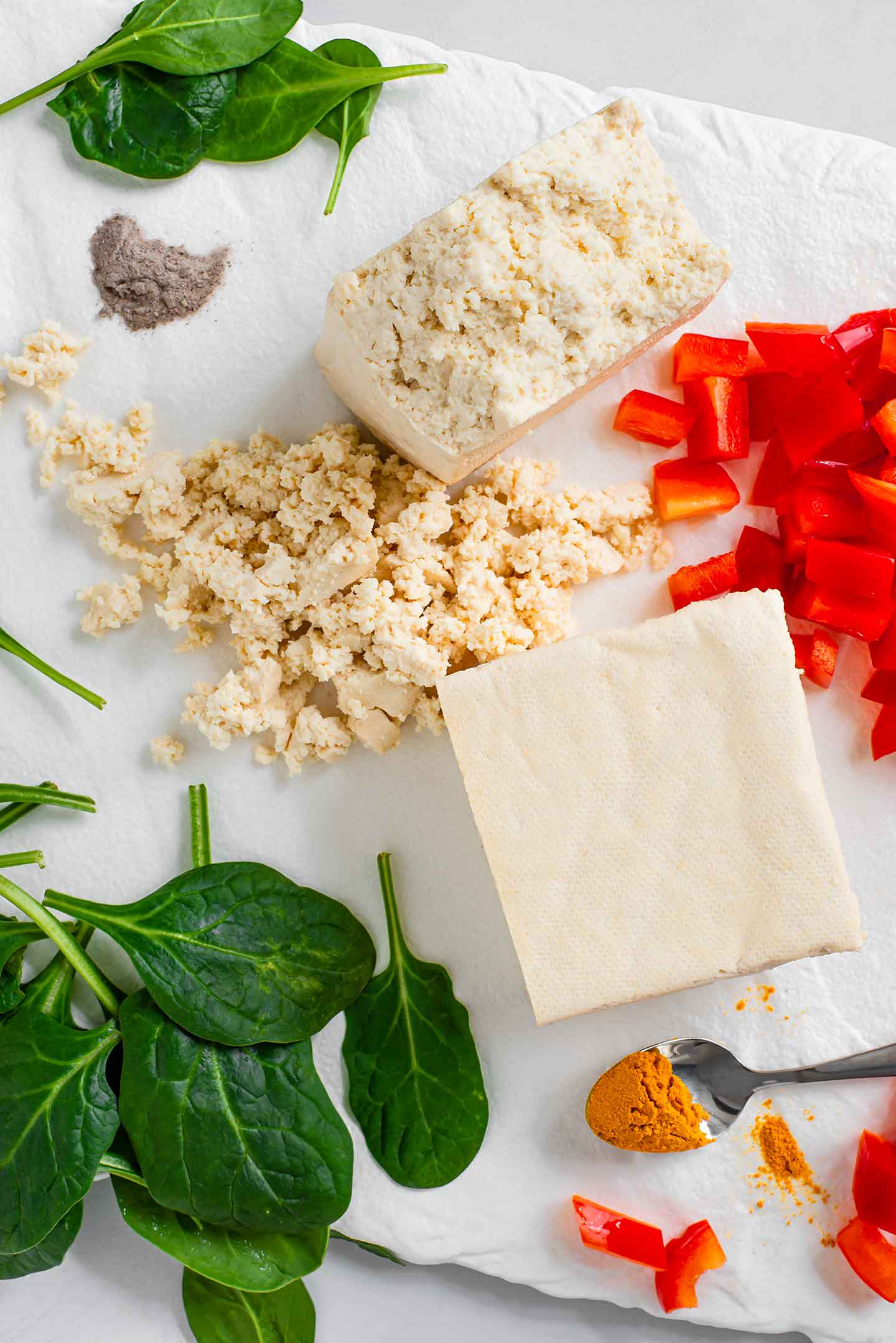 Top down view of quick, easy scramble ingredients. A block of firm tofu is cut in half with one part crumbled. Ground turmeric, black salt, spinach, and diced red pepper surround the tofu on a white tray.