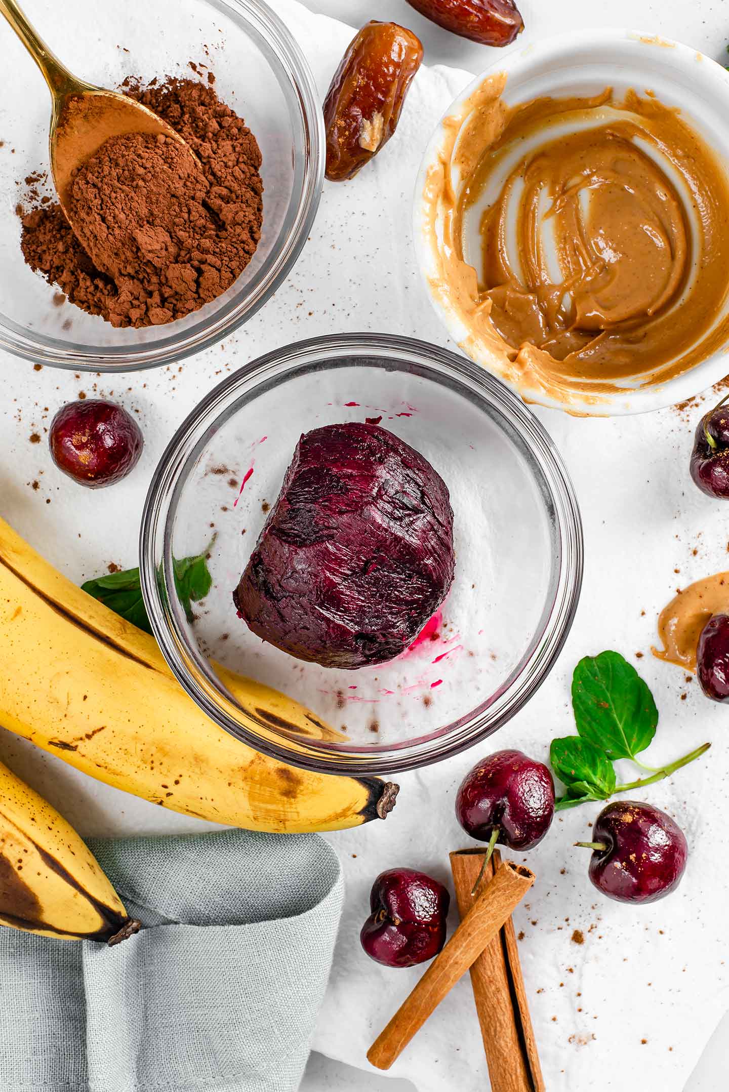 Top down view of a roasted beet in a glass dish with bananas, cocoa powder, peanut butter, cherries, dates, and cinnamon sticks scattered around.
