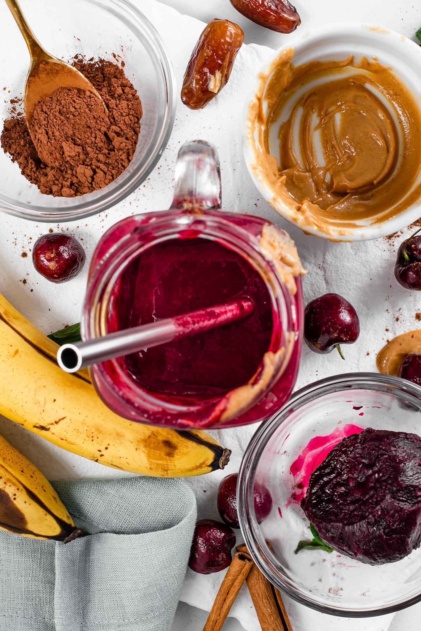 Top down view of a half drunk chocolate beet smoothie with a stainless steel straw. A roasted beet rests in a glass dish to the side. Bananas, cocoa powder, peanut butter, cherries, and dates are scattered around.