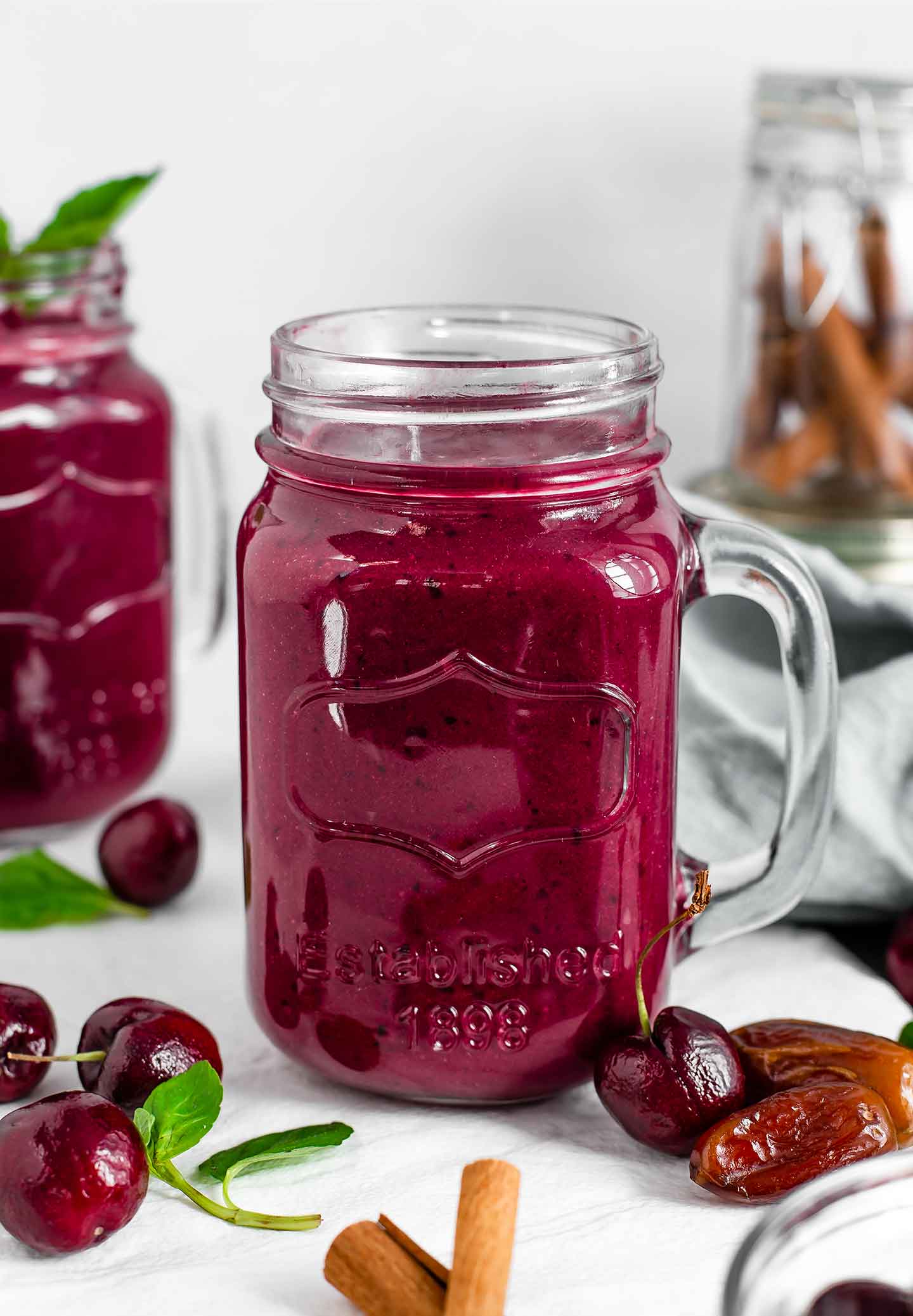 Side view of chocolate beet smoothie in a glass jar with cinnamon sticks, cherries, and dates scattered around. Another smoothie with a mint leaf stands in the background.