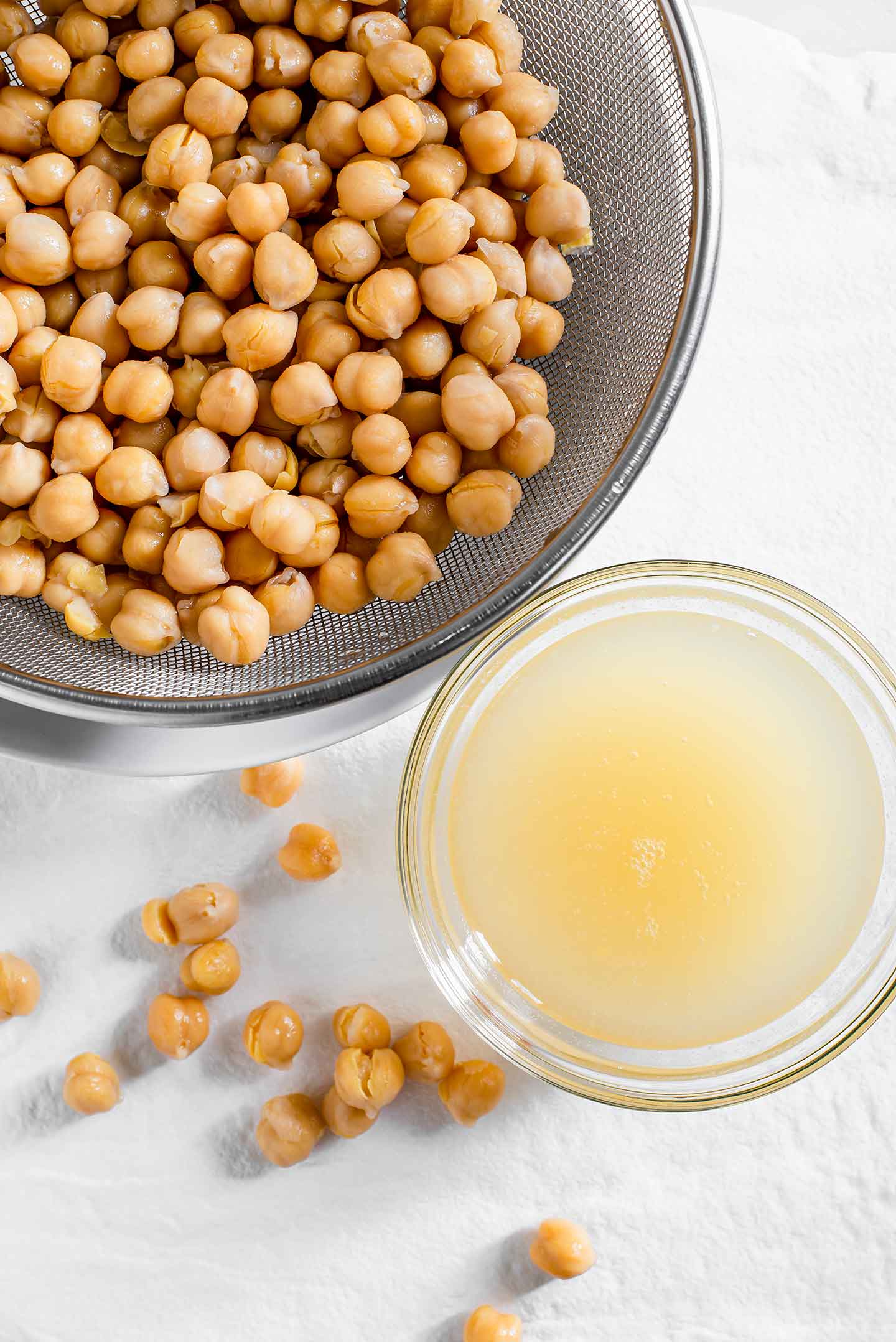 Top down view of chickpeas draining in a metal sieve with aquafaba reserved in a small bowl to the side.