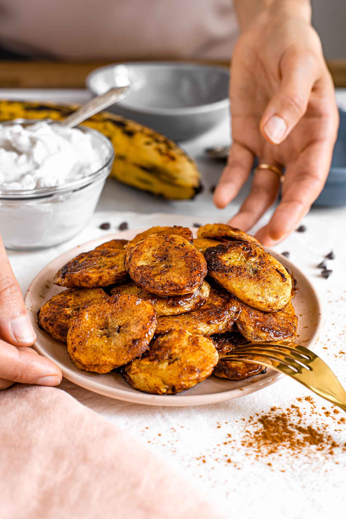 Side view of hands placing a plate of golden, crispy, caramelized plantains on a white tray with whipped cream in the background.