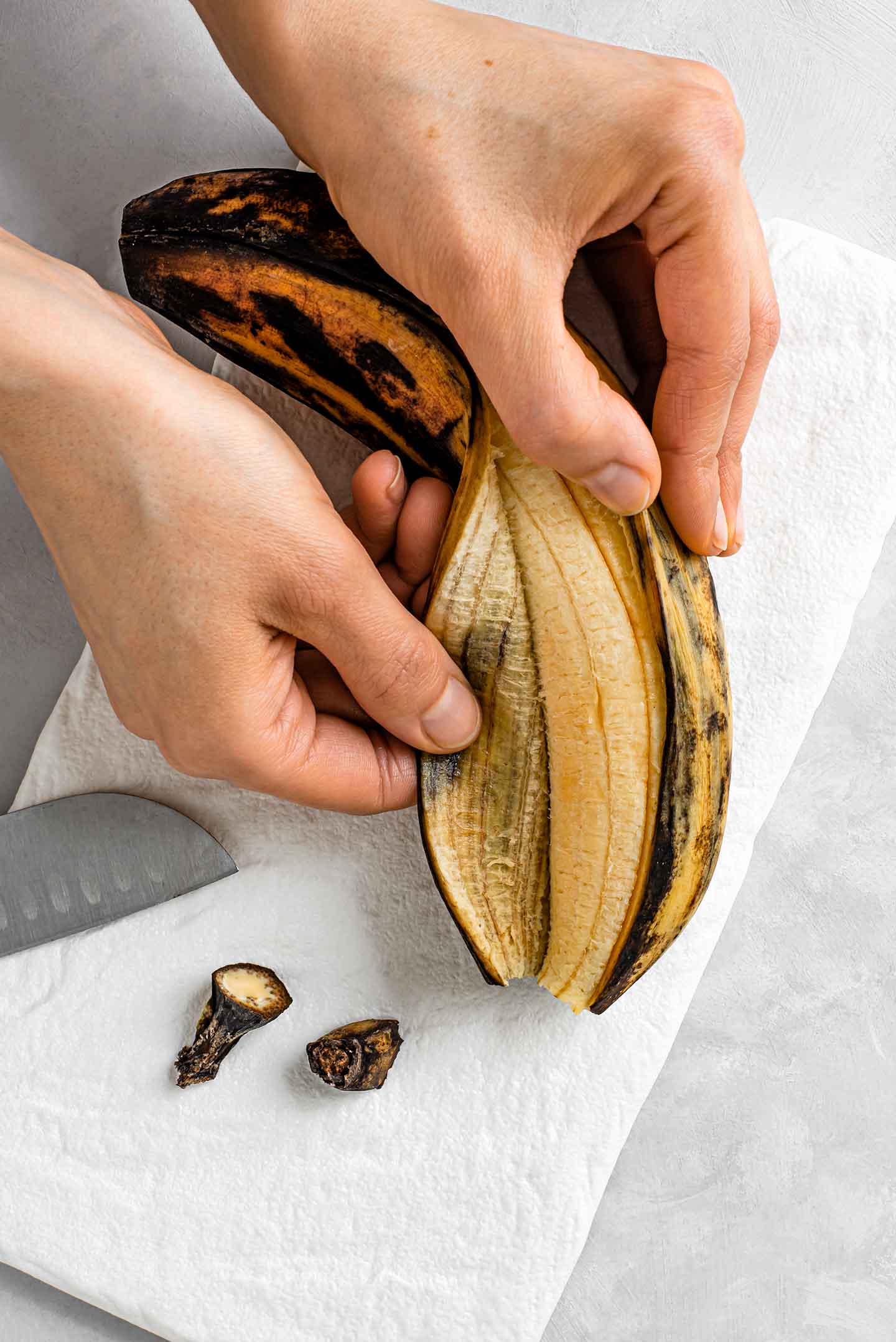 Top down view of two hands pulling the peel back from a plantain. The peel has been sliced down the length of the fruit and the two ends lay sliced off on a tray.