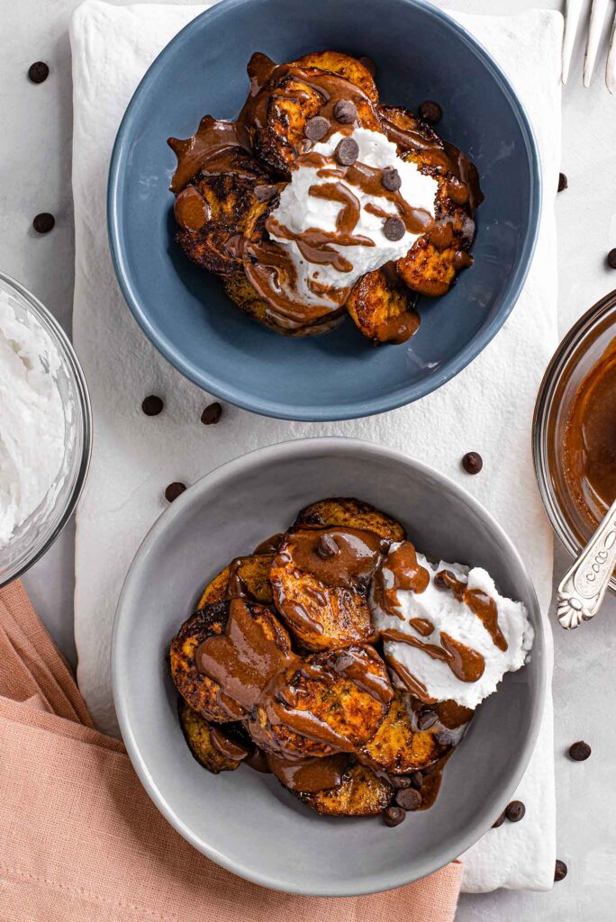 Top down view of two bowls of caramelized plantains with whipped cream, chocolate drizzle, and chocolate chips.