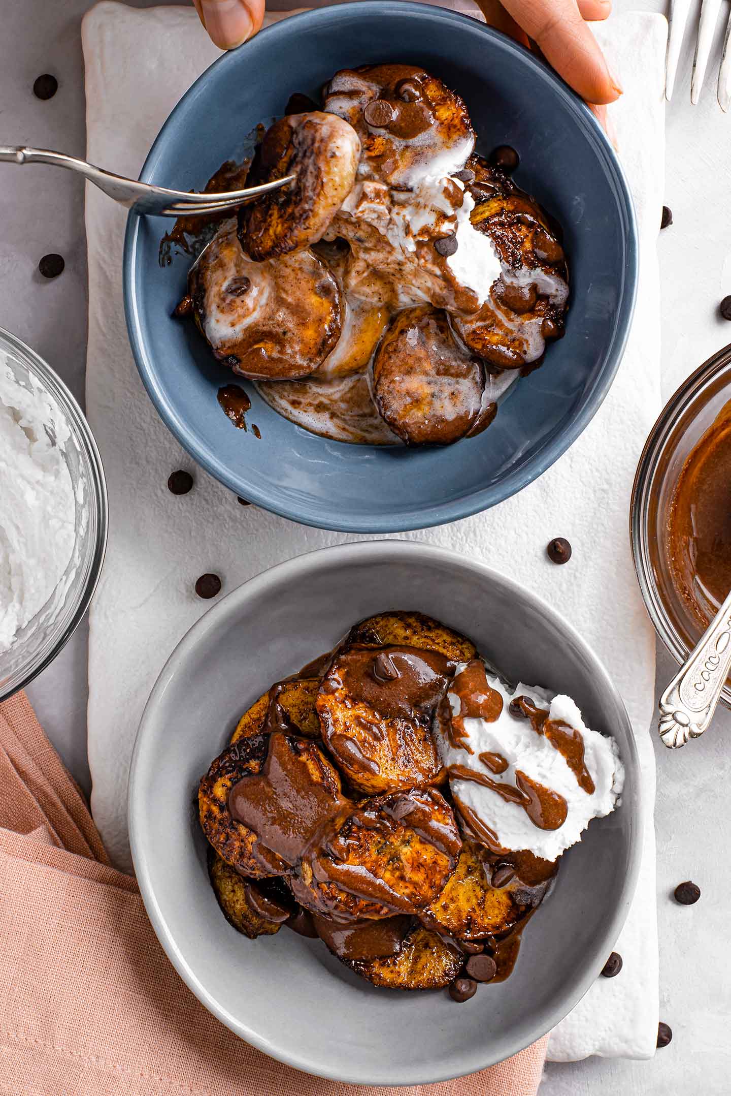 Top down view of two bowls of warm caramelized plantains. A hand uses a fork to pick up a slice from one bowl. The warm plantains have melted whipped cream and mix with chocolate syrup.