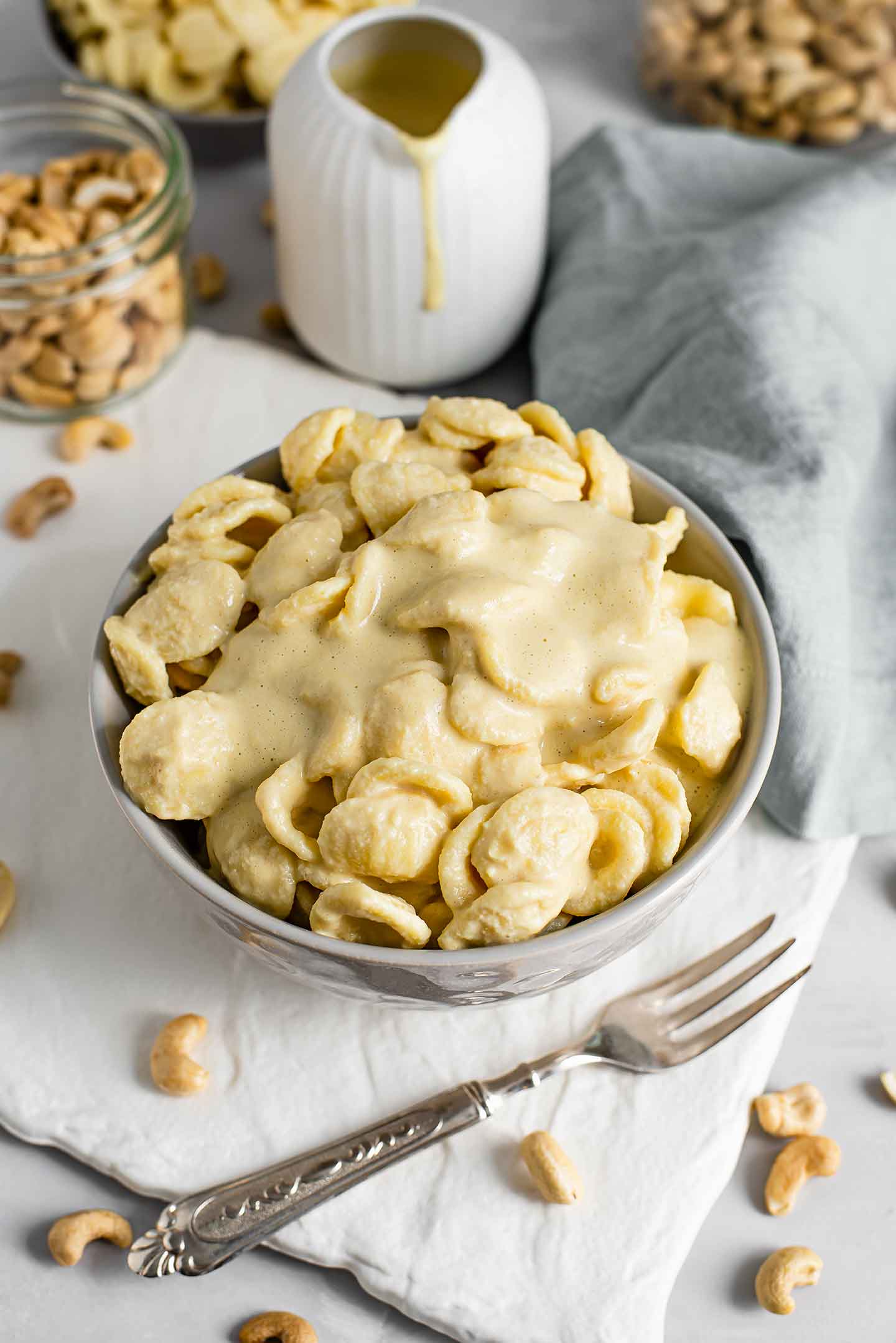 Top down side view of creamy vegan mac and cheese sauce on pasta shells. Extra sauce drips down a jar behind the bowl of pasta and raw cashews are scattered around the bowl.