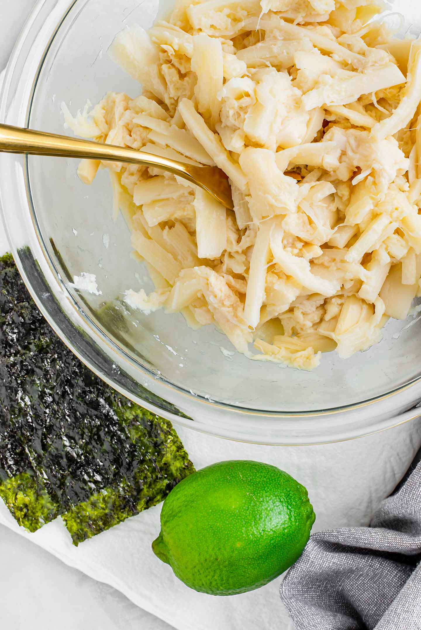Top down view of shredded hearts of palm in a glass bowl with a lime and seaweed snack sheets nearby.