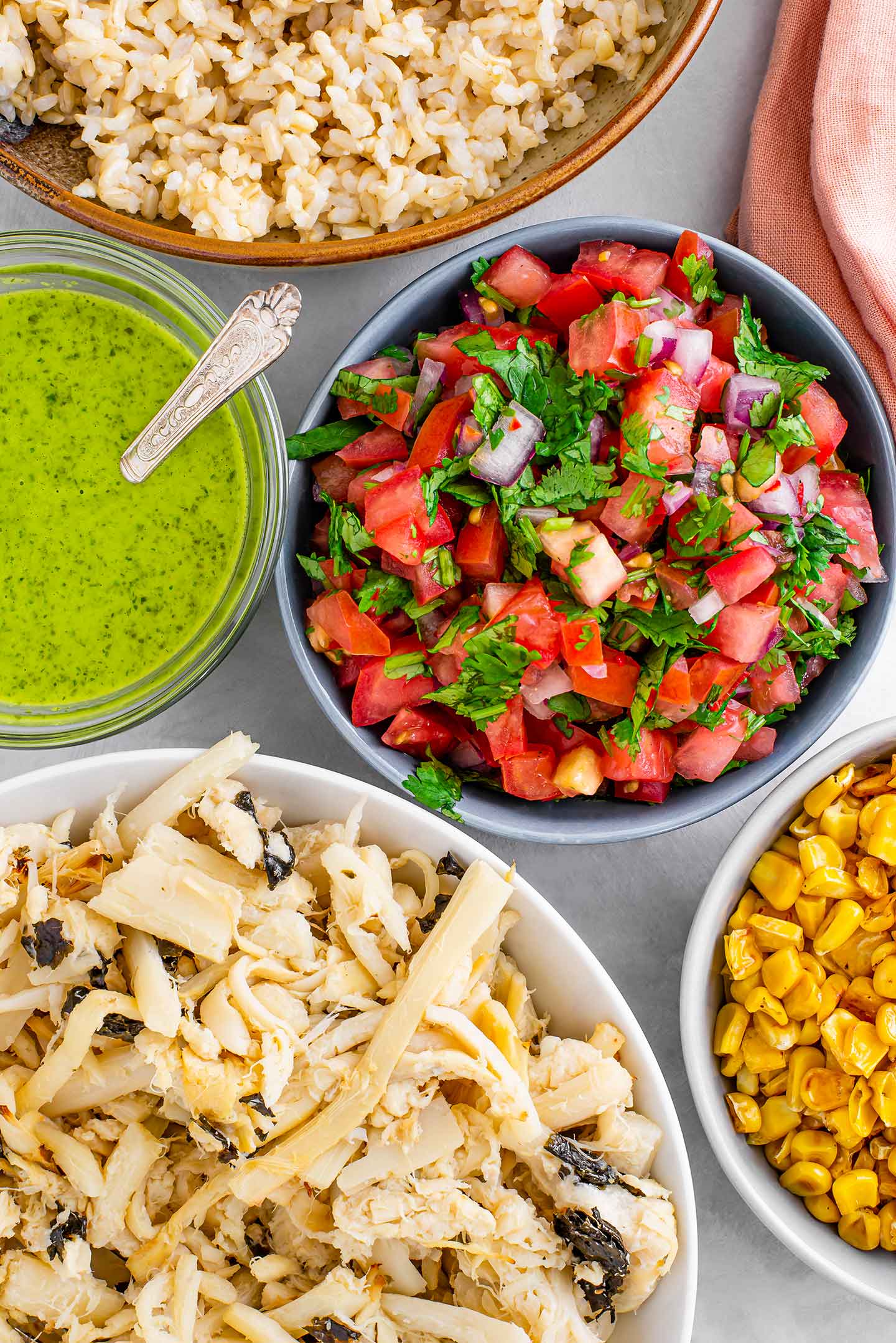Top down view of easy "fish" taco bowl ingredients. Salsa fresca fills a small bowl, bright green cilantro lime vinaigrette is in a small dish with a spoon, the baked hearts of palm, charred corn, and brown rice surround.