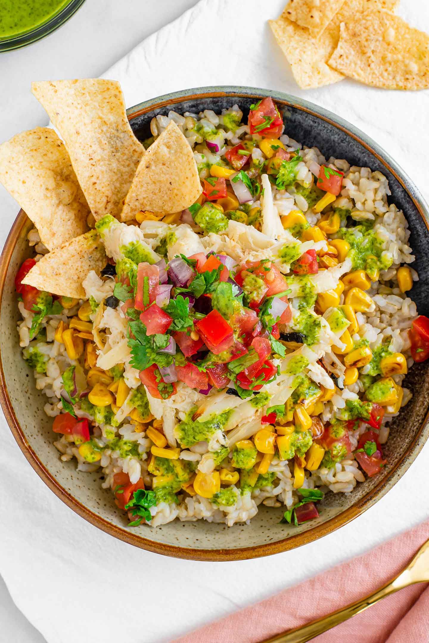 Top down view of the colourful completed bowl. Brown rice, charred corn, hearts of palm and salsa fresca are all piled on one another. Cilantro lime vinaigrette is drizzled over and tortilla chips garnish one side.