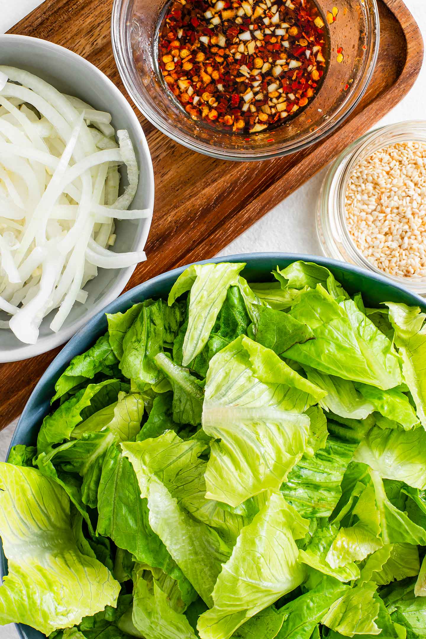 Top down view of the prepared ingredients. Torn lettuce, sliced onion, a finished dressing, and toasted sesame seeds.