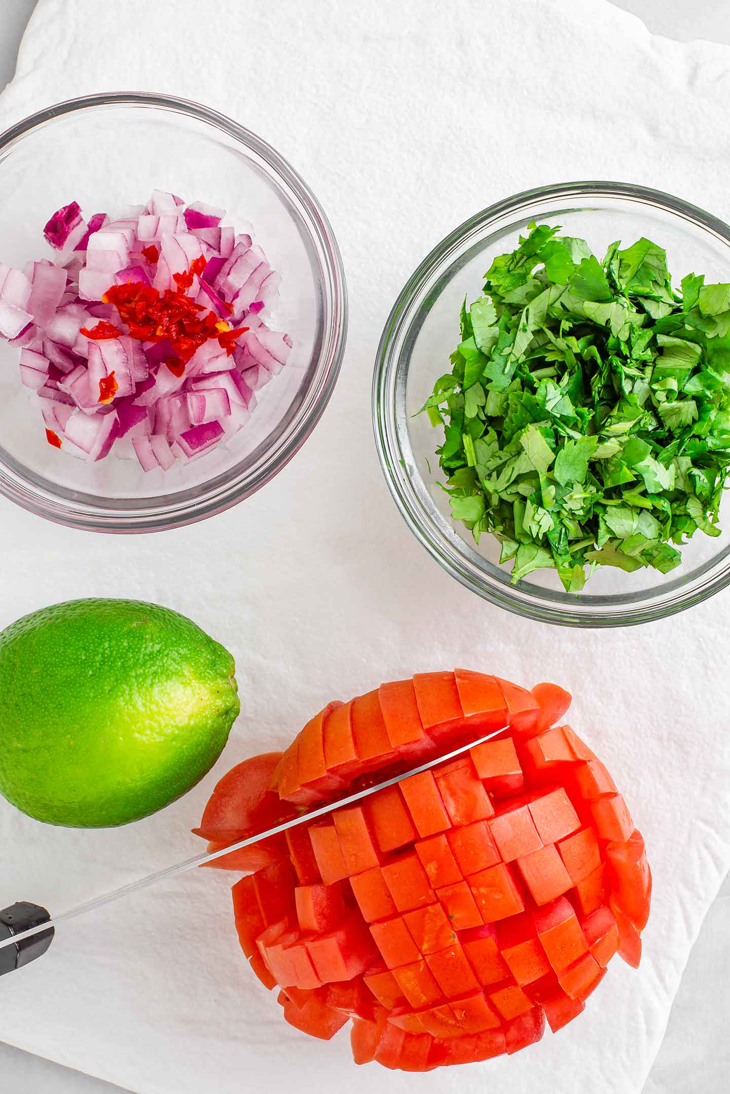 Top down view of a tomato still intact but sliced in a checkered pattern. A knife rests in one of the cuts and diced red onion, chopped cilantro, and a fresh lime surround the tomato.