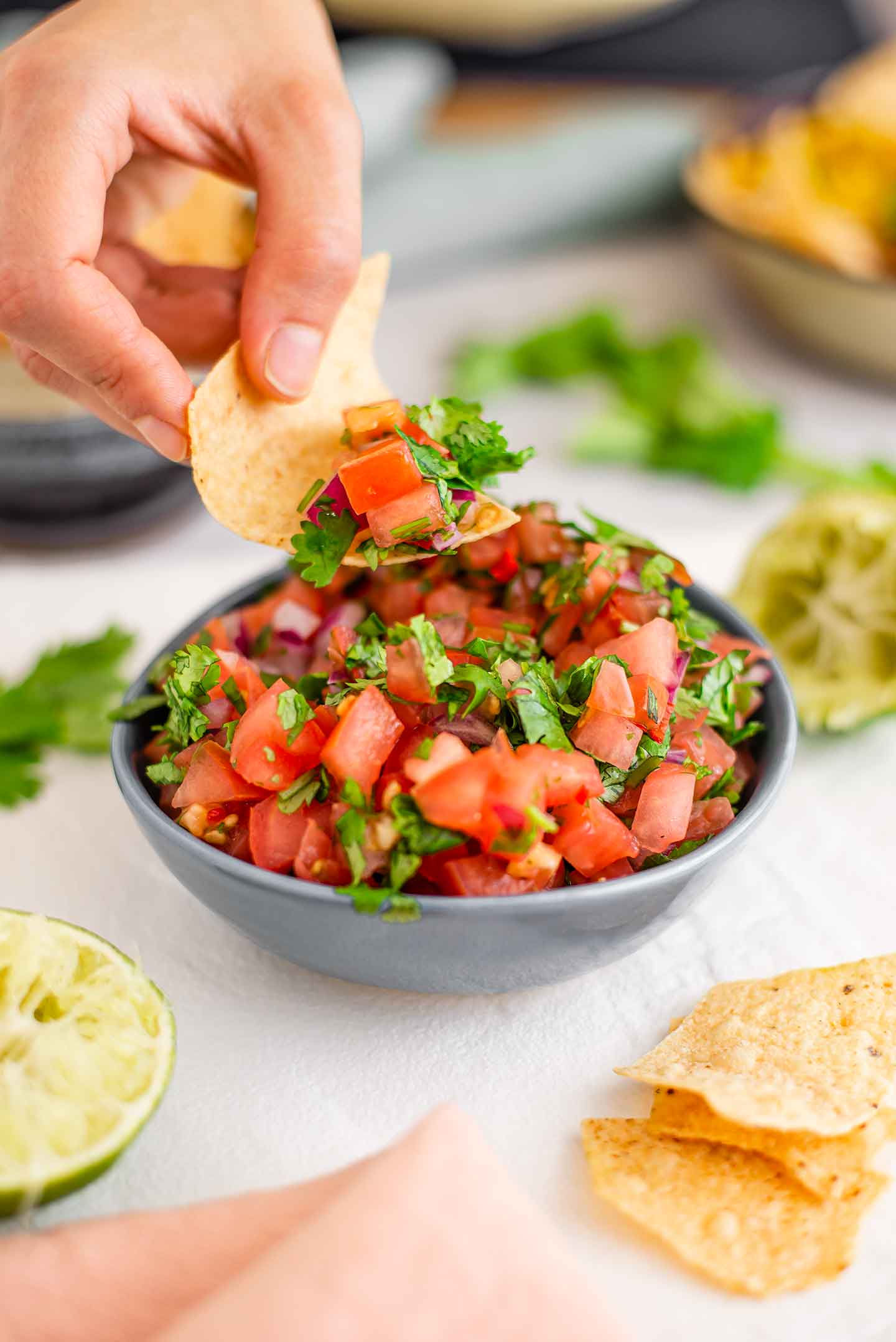 Salsa fresca with diced tomato, red onion, and cilantro fills a small dish. A hand scoops a tortilla chip into the chunky salsa.