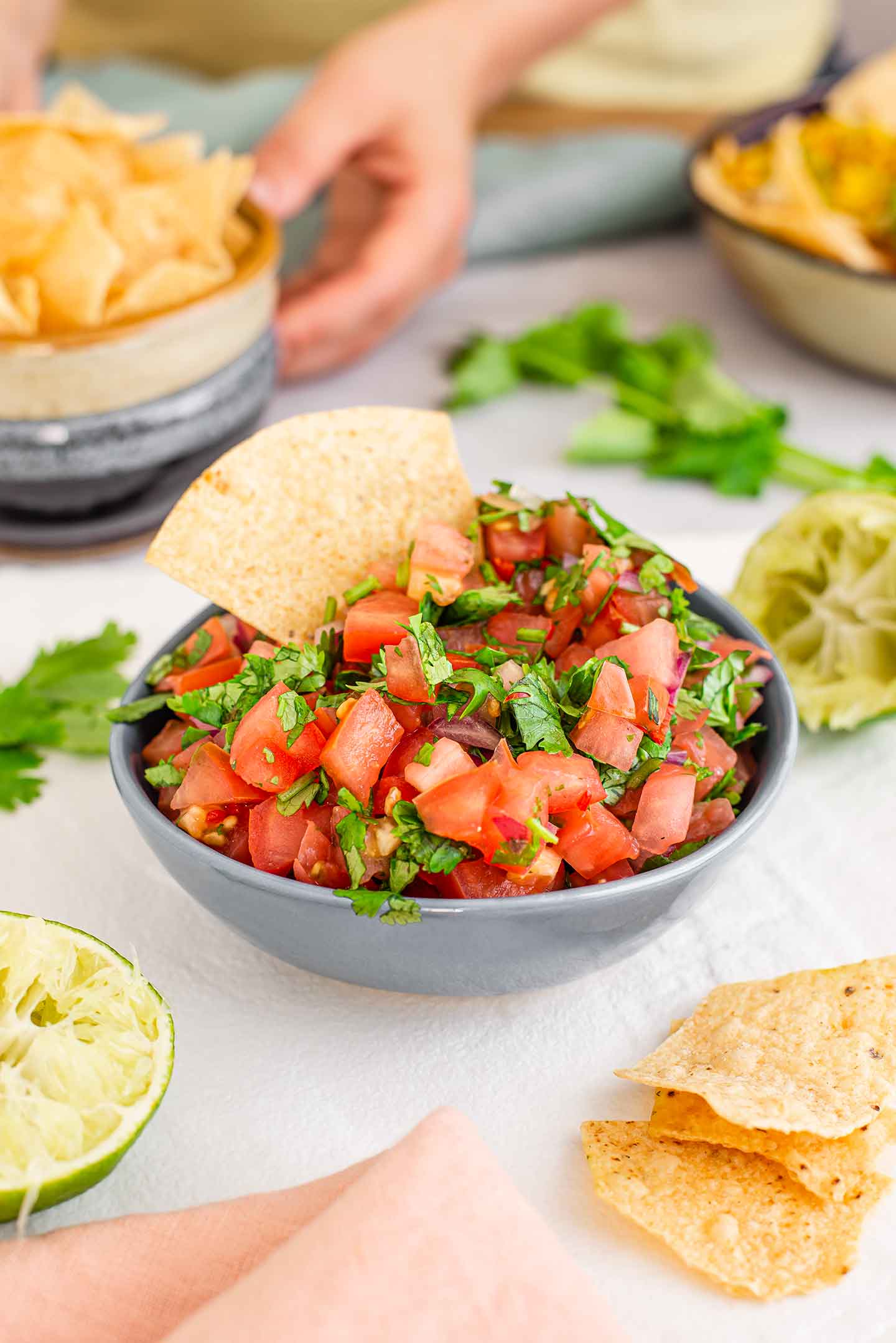 Salsa fresca fills a small dish. A tortilla chip is placed inside the bowl of chunky salsa. Juiced halves of lime, cilantro, and more tortilla chips surround the colourful salsa.