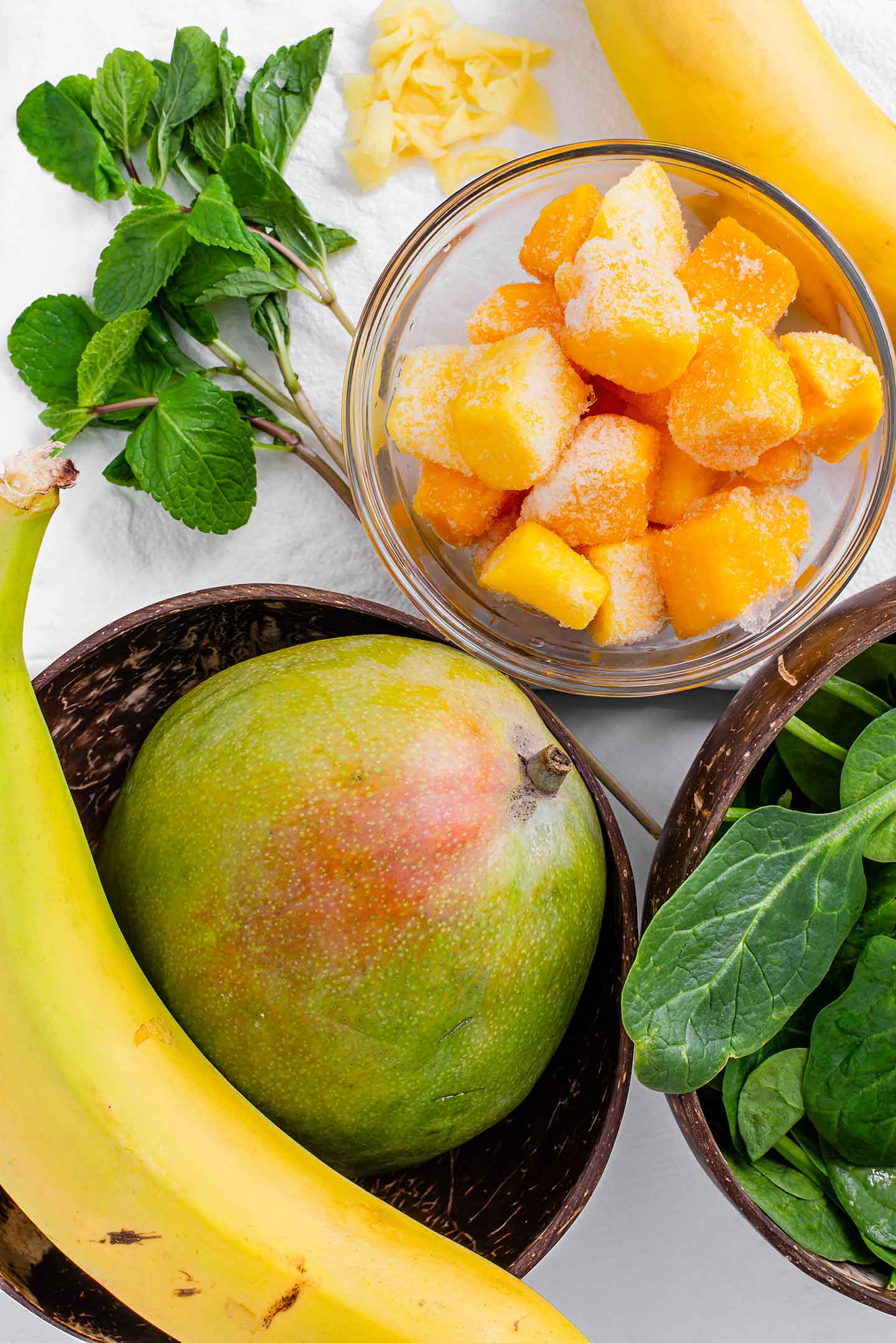 Top down view of a mango, banana, mint, diced frozen mango and spinach.