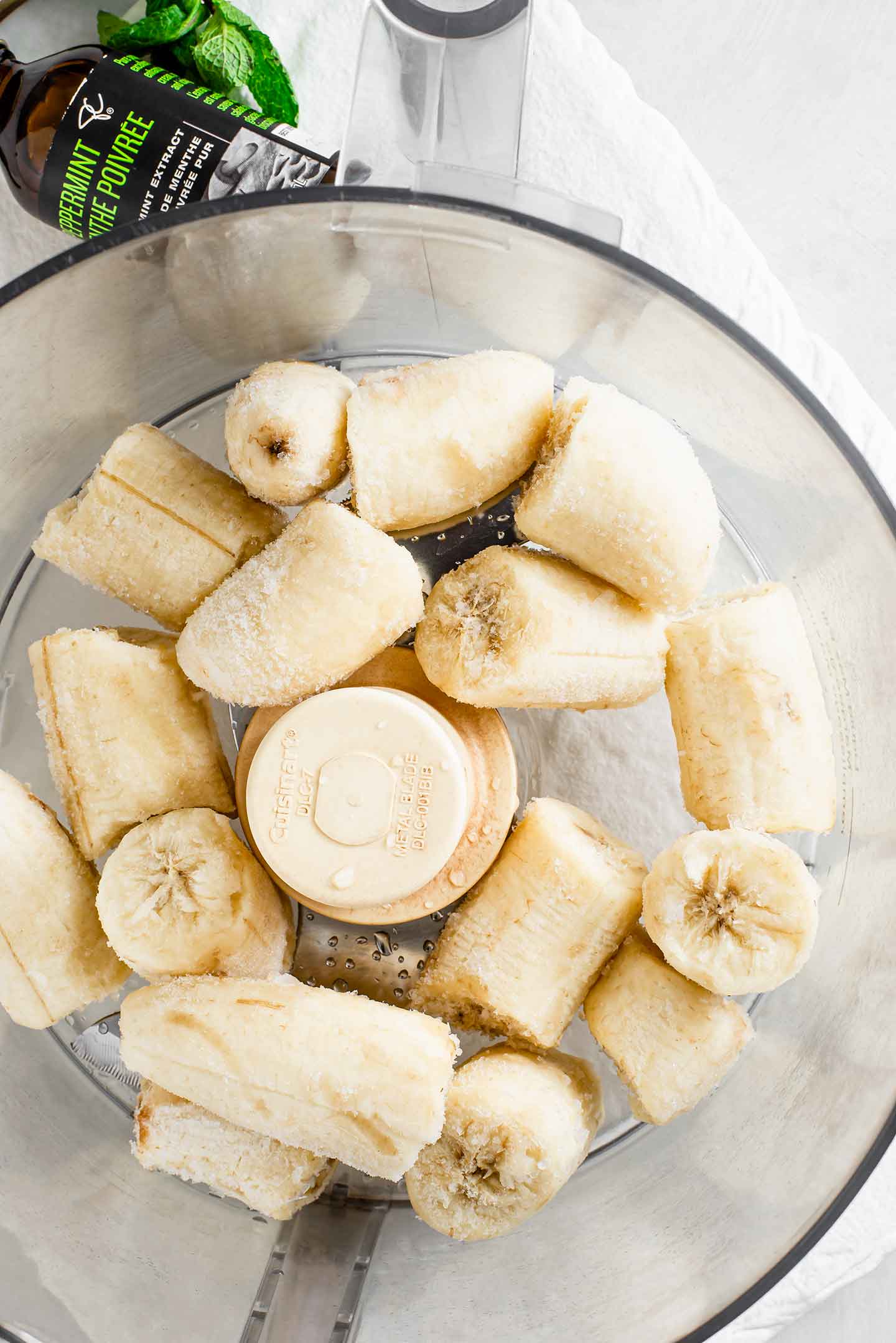 Top down view of frozen banana chunks in a food processor. A jar of pure peppermint extract rests on a tray.