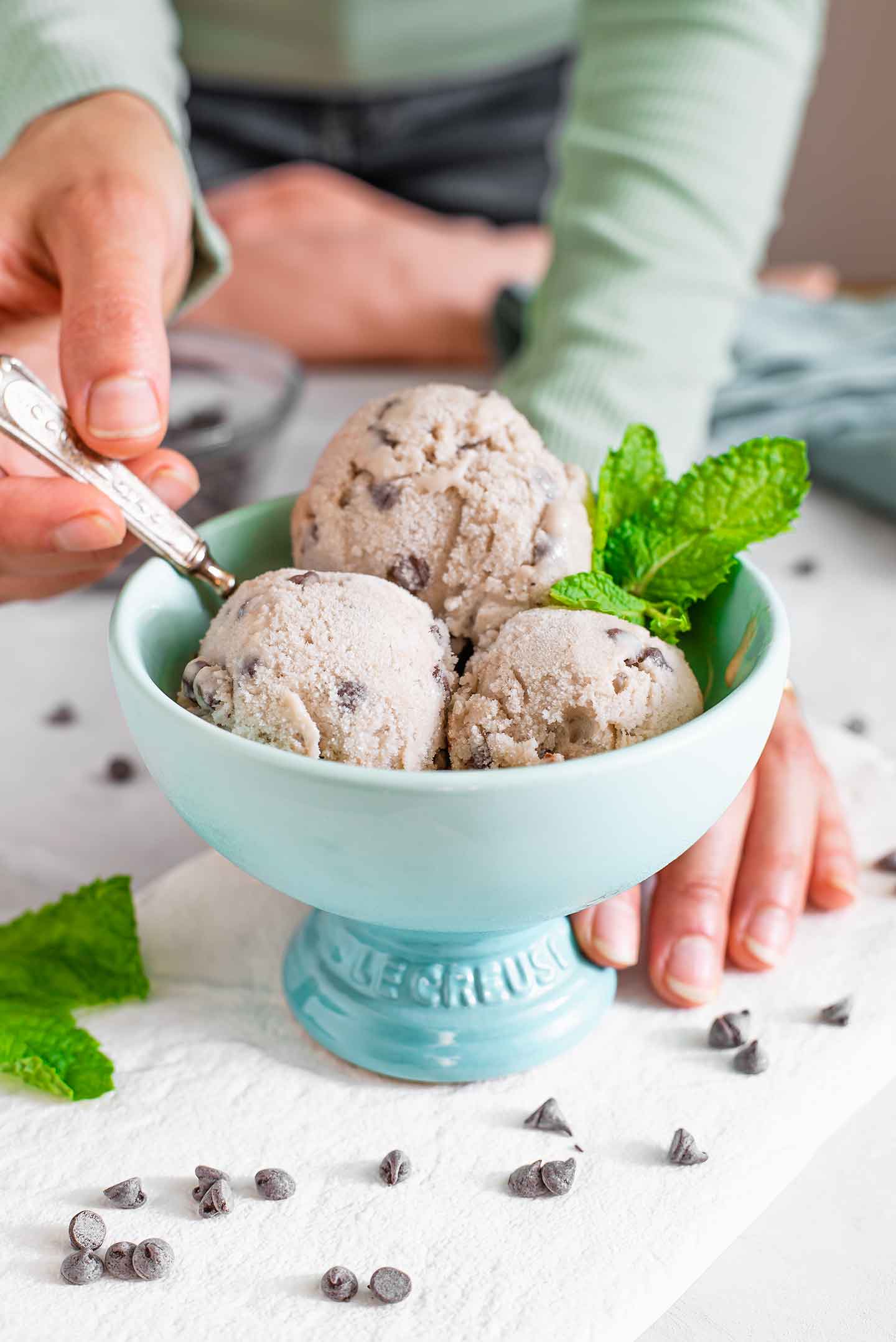 Side view of three scoops of mint chip nice cream in a sorbet bowl. A hand dips a spoon into the creamy nice cream and chocolate chips are scattered on the tray below.