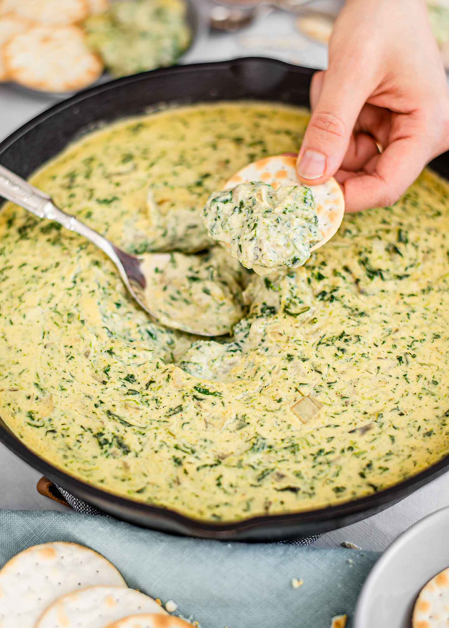 Side view of a hand holding a cracker with a scoop of spinach artichoke bean dip over the warm skillet of dip.