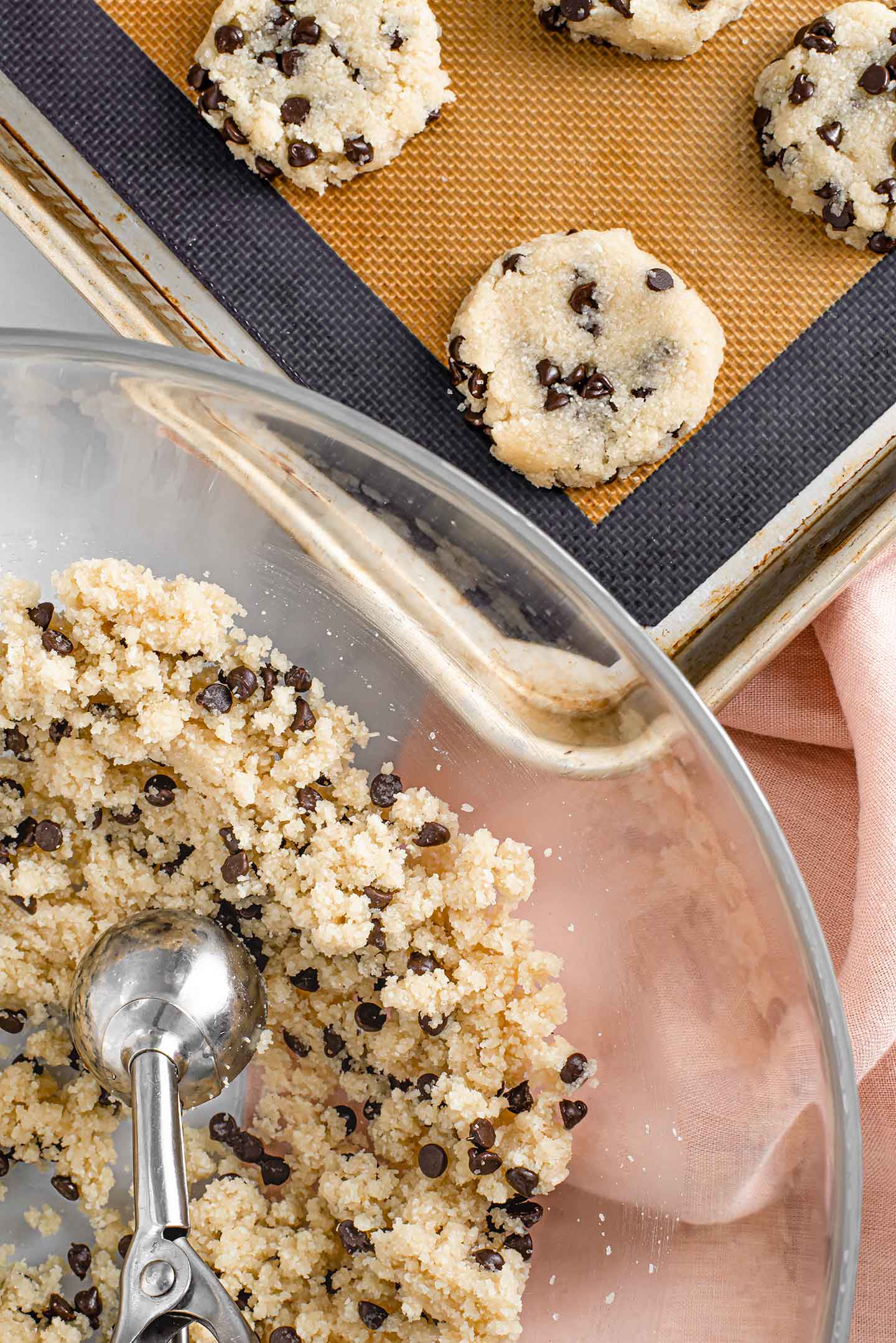 Top down view of raw chocolate chip cookies on a baking sheet with the rest of the chocolate chip cookie dough batter in a glass mixing bowl.