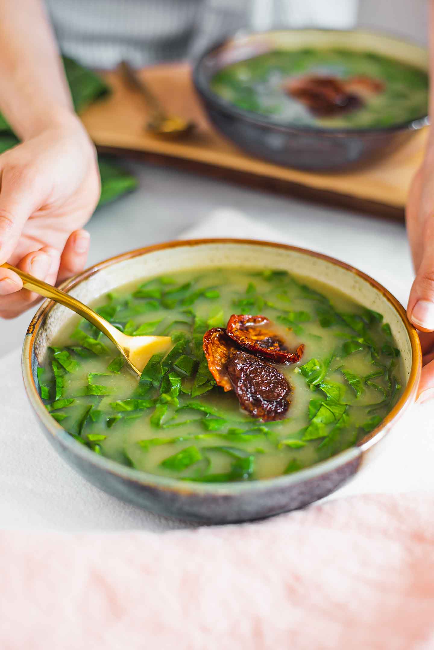 Side view of a hand holding a spoon and dipping it into the caldo verde. Another bowl is in the background and both are topped with a few pieces of smoky sun-dried tomato.
